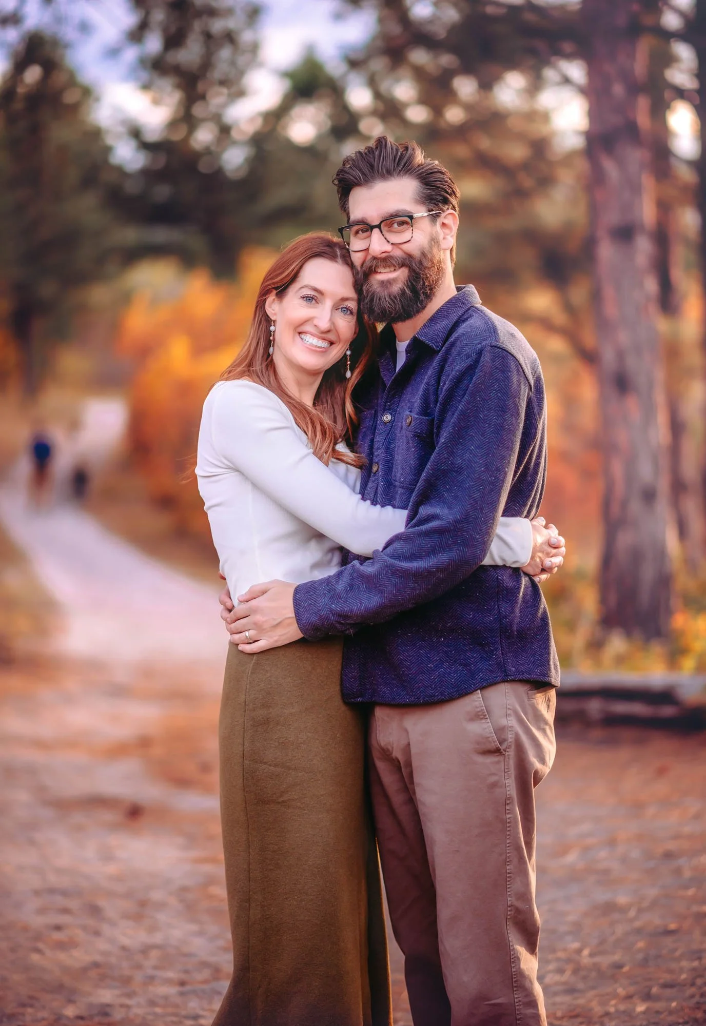 A happy couple hugging outdoors in autumn with trees and falling leaves in the background.