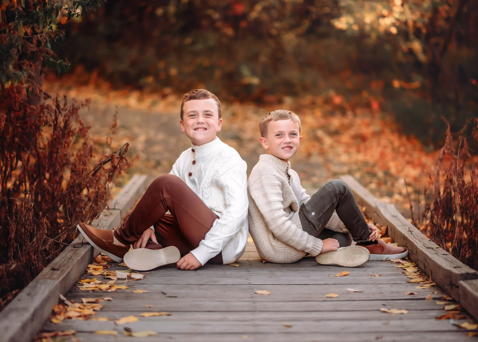 Two young boys sitting back-to-back on a wooden bridge surrounded by fall foliage with autumn leaves on the ground.
