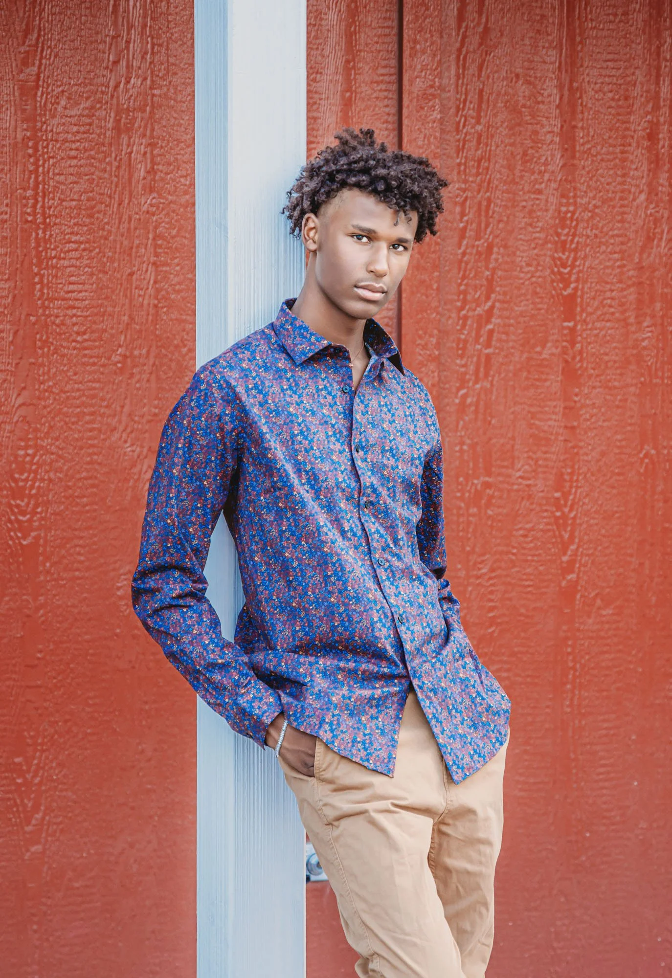 A young man with curly dark hair wearing a blue patterned shirt and beige pants, leaning against a blue metal pole in front of a red wooden wall.