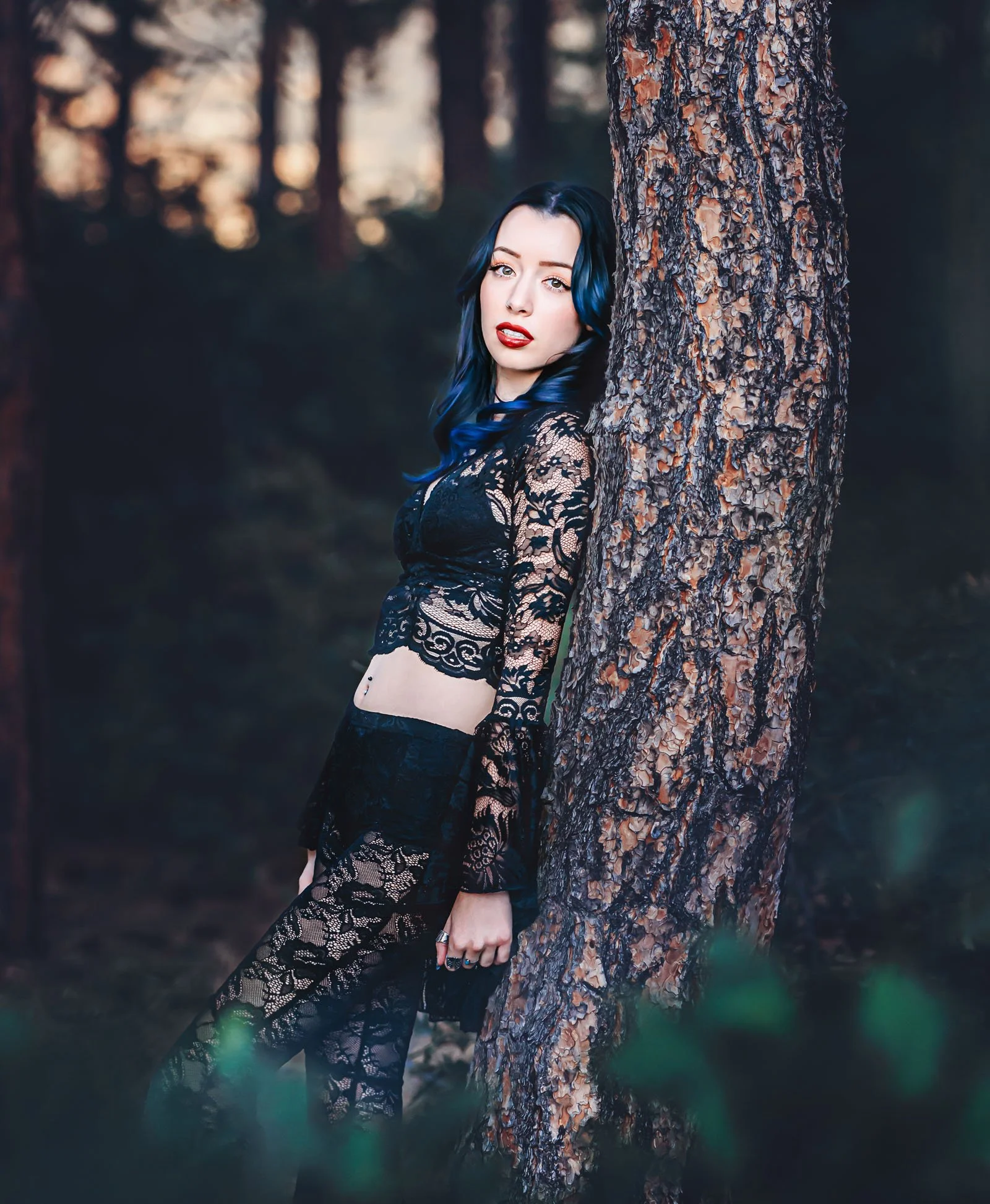 A woman with blue and black hair leaning against a tree trunk in a forest, wearing a black lace dress.