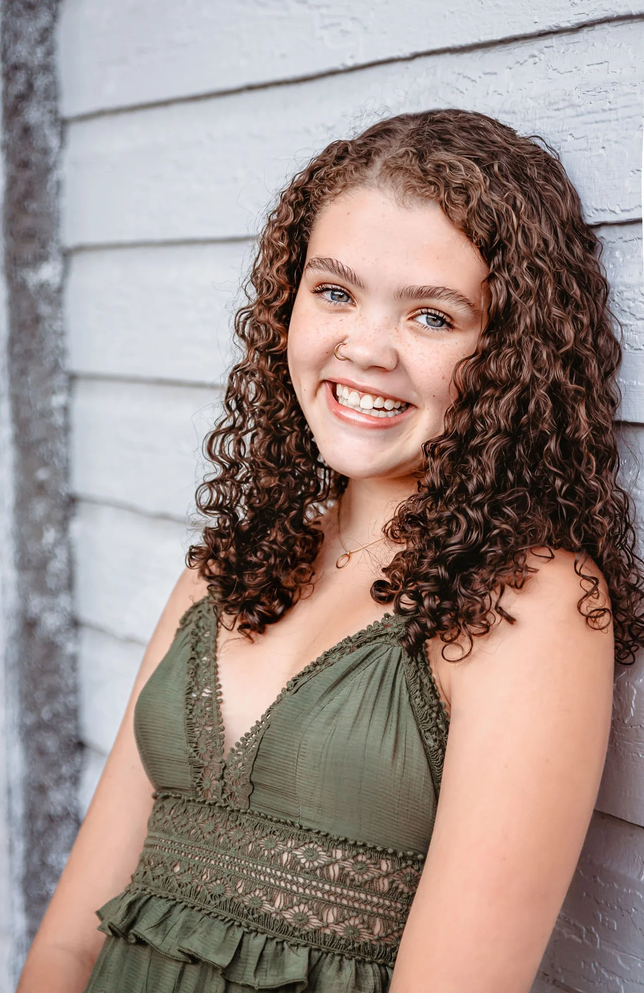 A young woman with curly brown hair, blue eyes, and a nose ring smiling at the camera. She is wearing a green lace-trimmed top and standing in front of a white wooden wall.