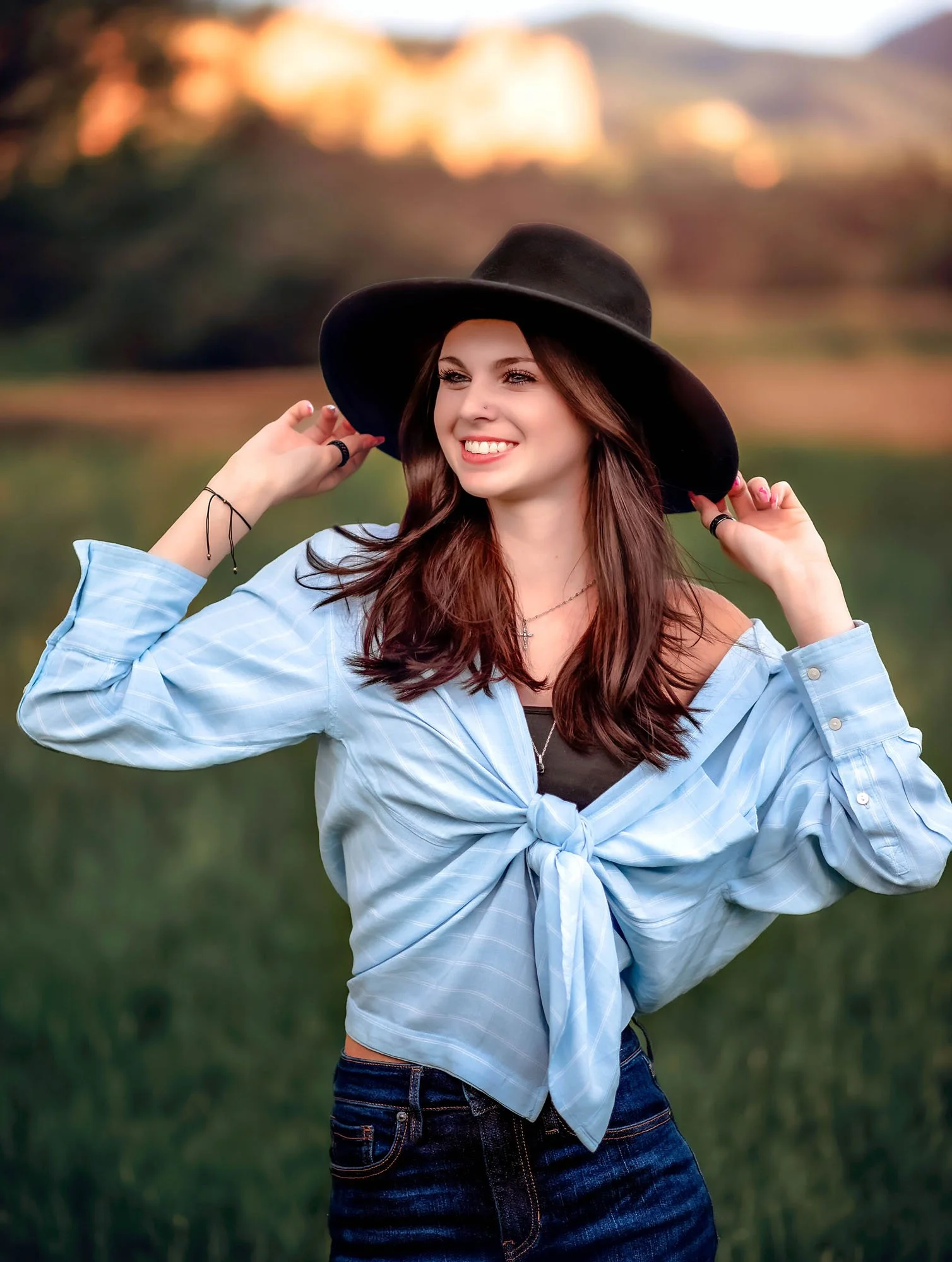 A young woman with long brown hair smiling while adjusting her black wide-brimmed hat outdoors during sunset, wearing a light blue tied-up shirt over a black top and dark blue jeans, with a blurred natural landscape in the background.
