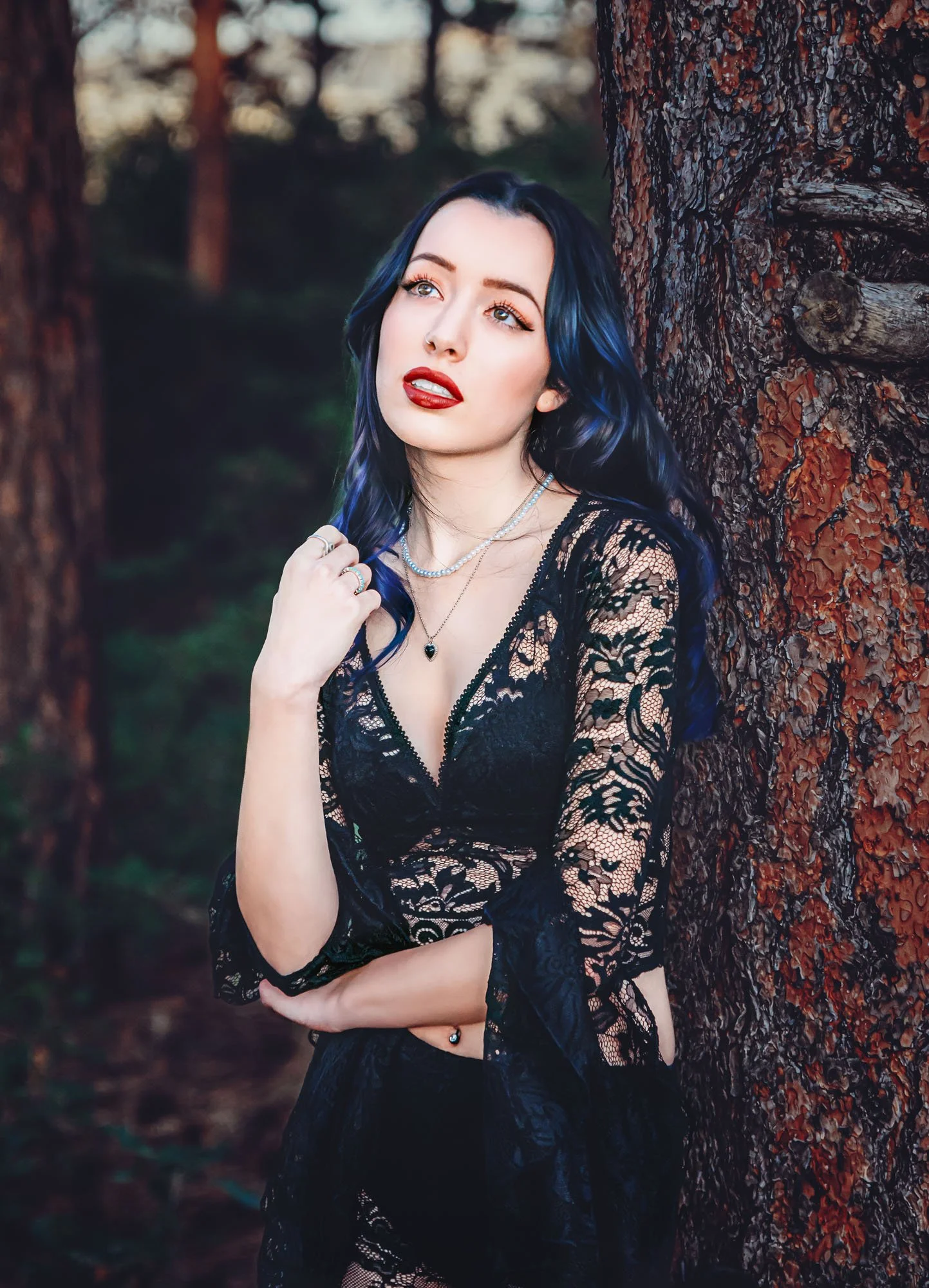 A young woman with blue and black hair, wearing a black lace dress and jewelry, leaning against a tree in a forest during sunset.