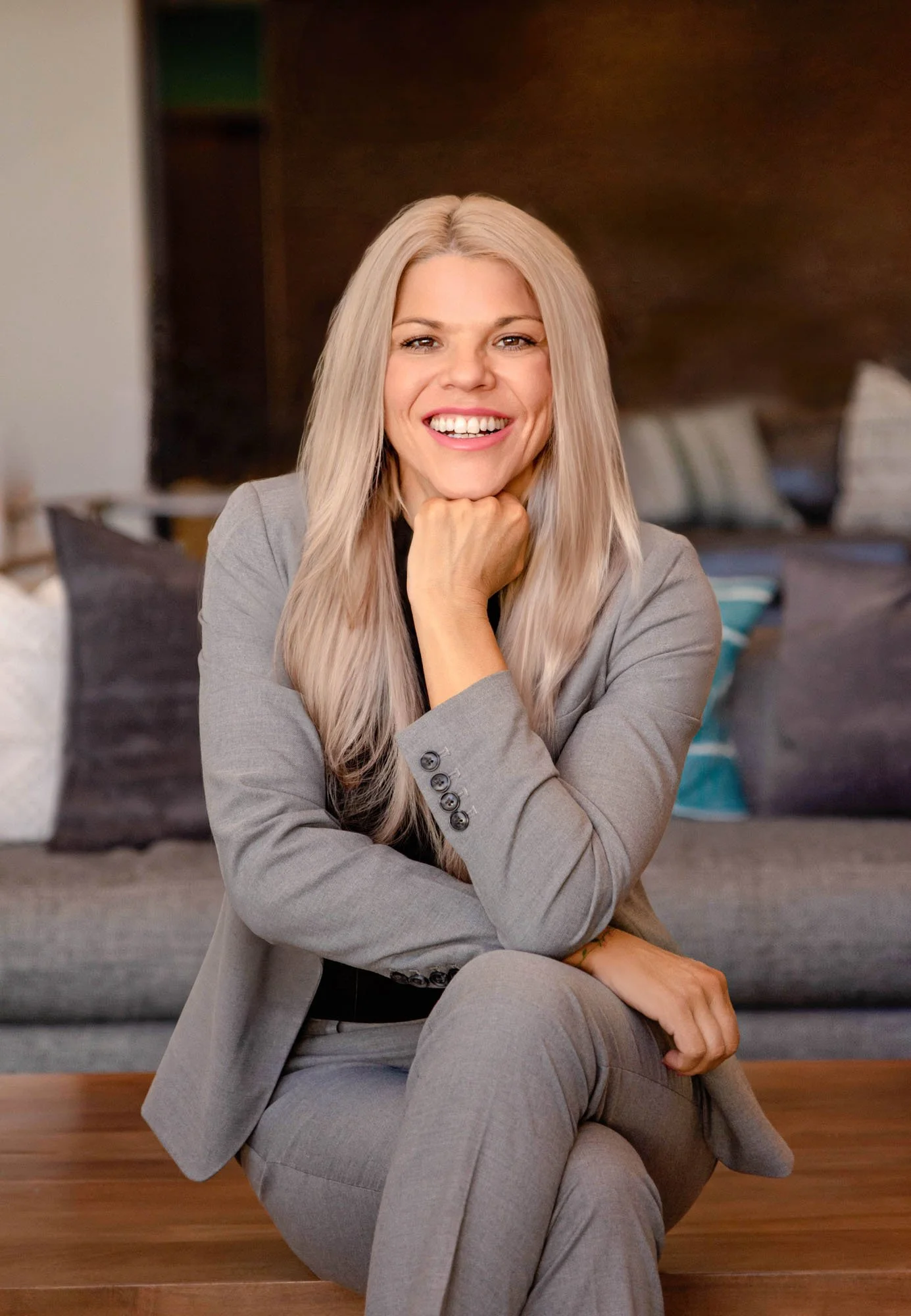 A woman with long blond hair smiling and sitting with her chin resting on her hand in a gray suit, with a blurred background of a living room.