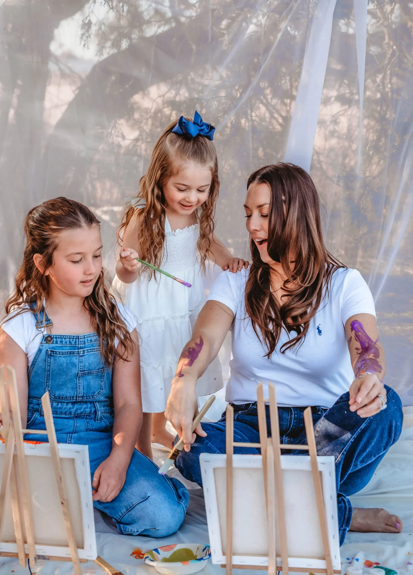 A woman and two girls painting on small canvases outdoors during daytime. The woman has long brown hair and is wearing a white shirt and jeans. One girl has long brown hair, a blue bow, and a white dress. The other girl has long brown hair, and is we