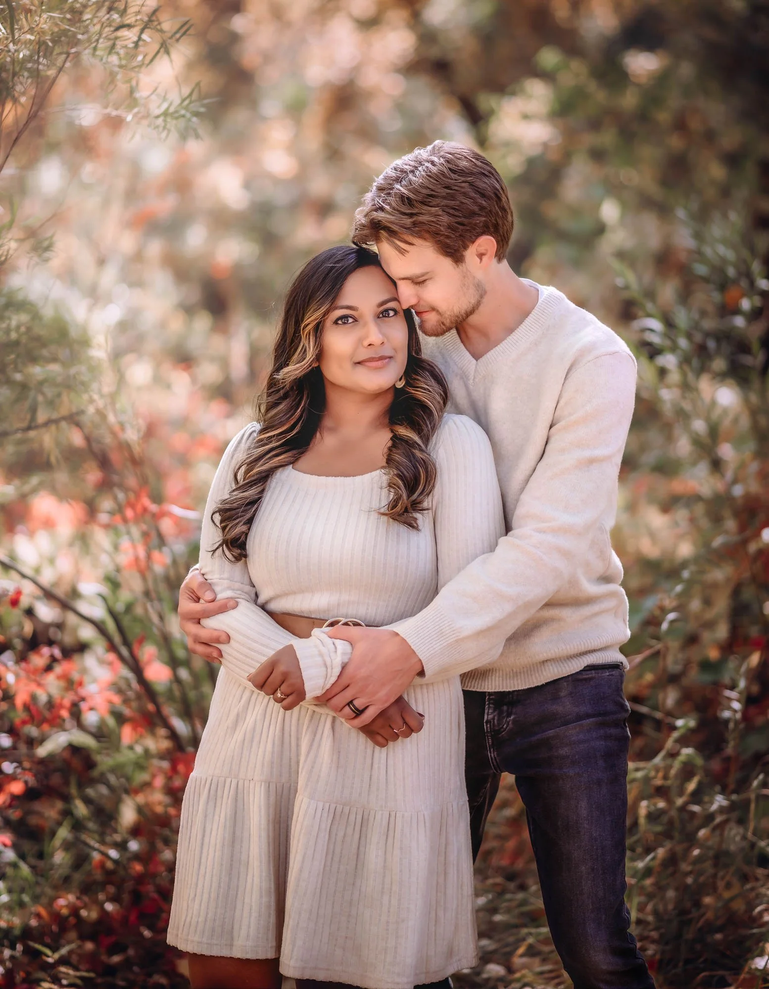 A young couple standing outdoors in autumn, closely embracing with the man gently resting his forehead on the woman's temple, surrounded by fall foliage.