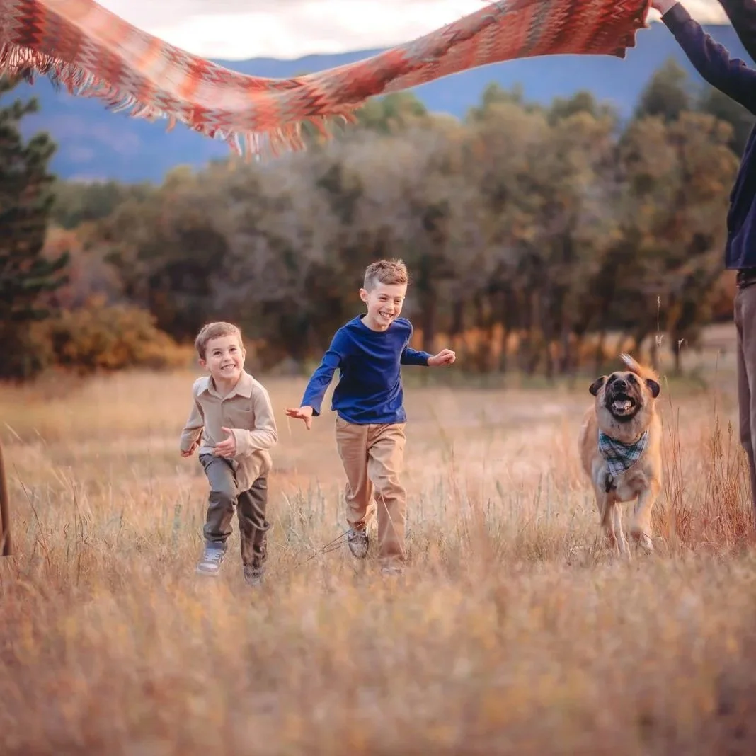 Two children running with a dog in a field during autumn, with mountains and trees in the background.