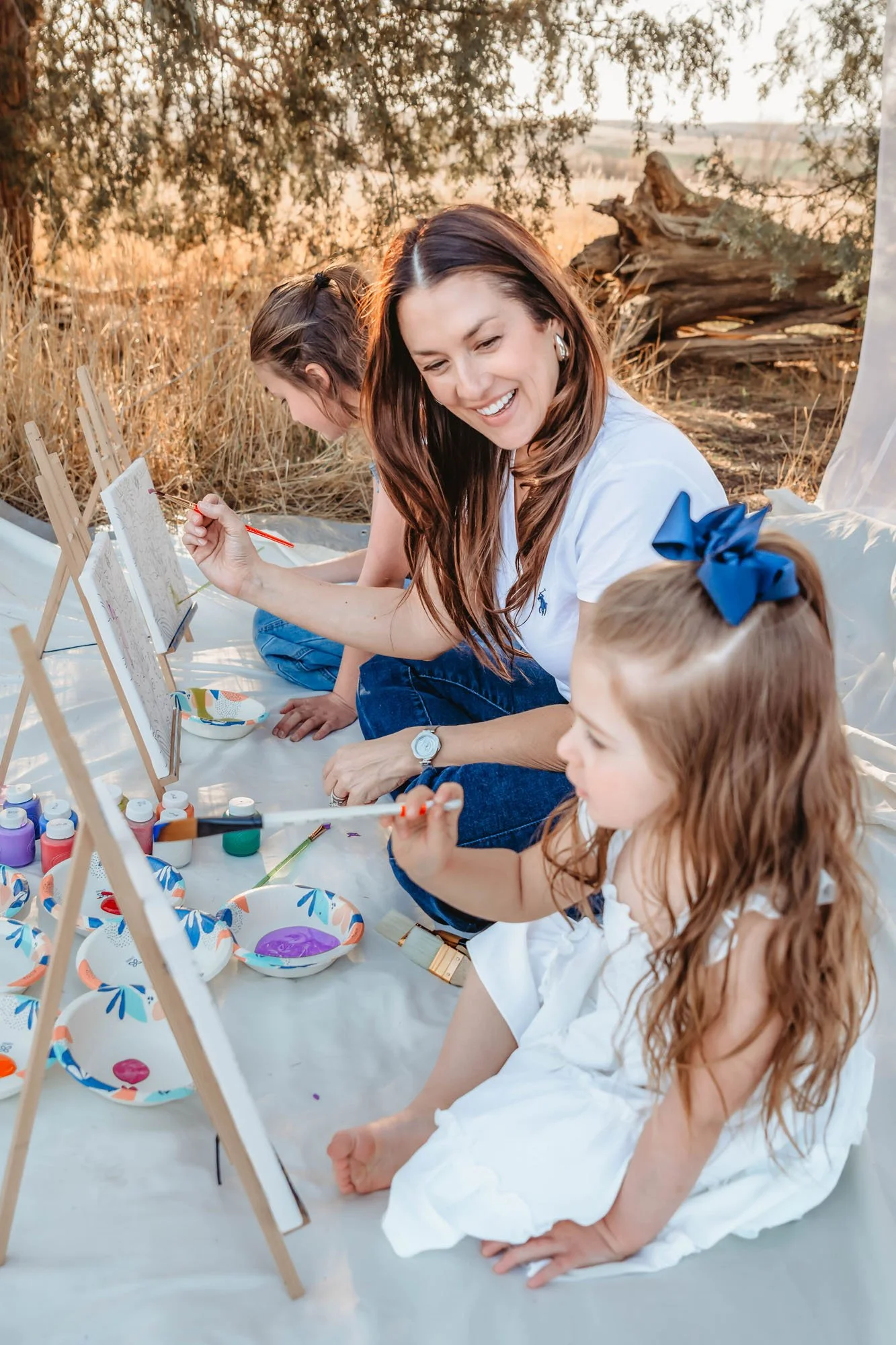 A woman and a young girl painting on canvases outdoors during sunset, surrounded by art supplies and decorative bowls, sitting on a white sheet with a tree and wooden log in the background.