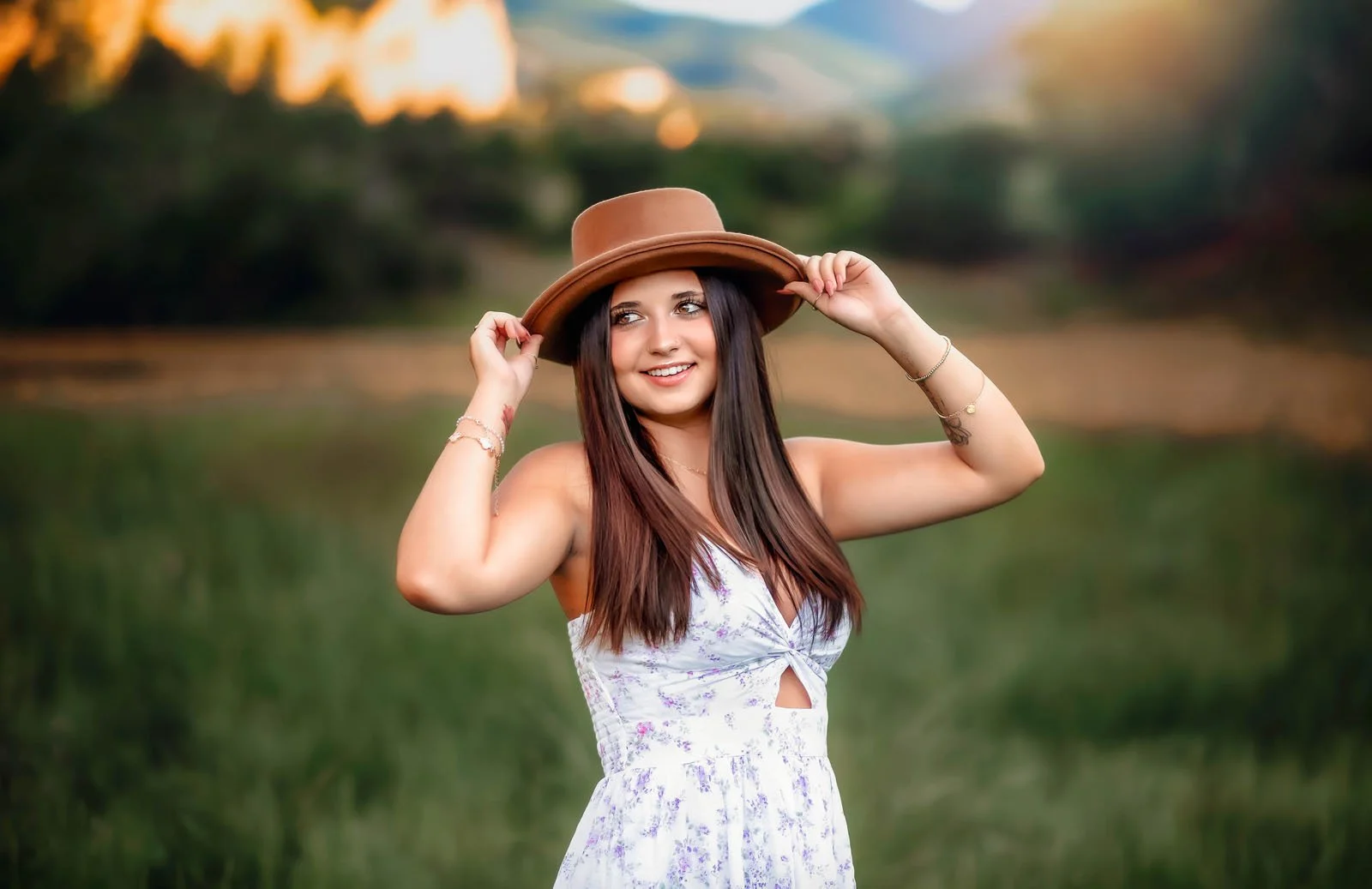 A smiling young woman in a white floral dress holds a brown hat on her head, standing outdoors in a natural setting during sunset.