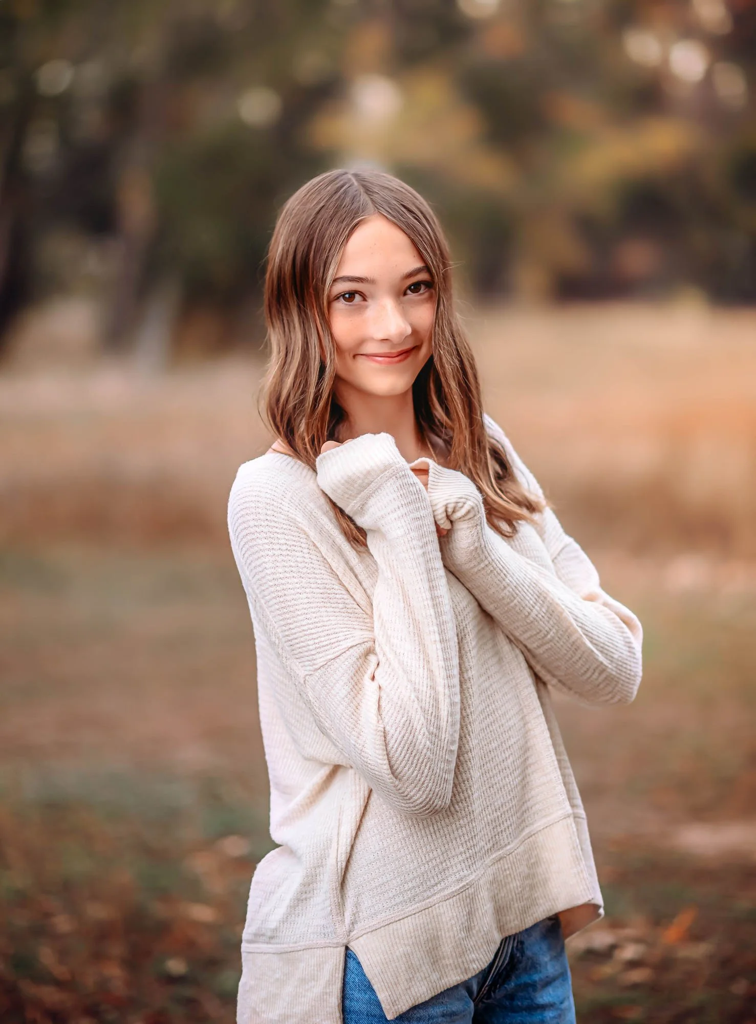 A young girl with long brown hair smiling outdoors during autumn, wearing a cream-colored sweater and blue jeans, with trees and fall leaves in the background.