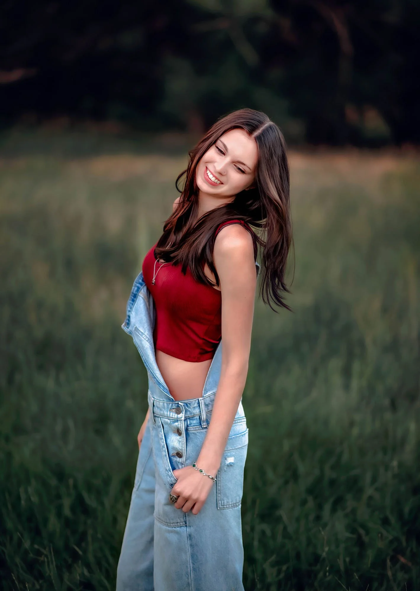 A young woman with long dark hair smiling and looking away, standing outdoors in a grassy area with trees in the background, wearing a red sleeveless top, light blue denim overalls, and jewelry.