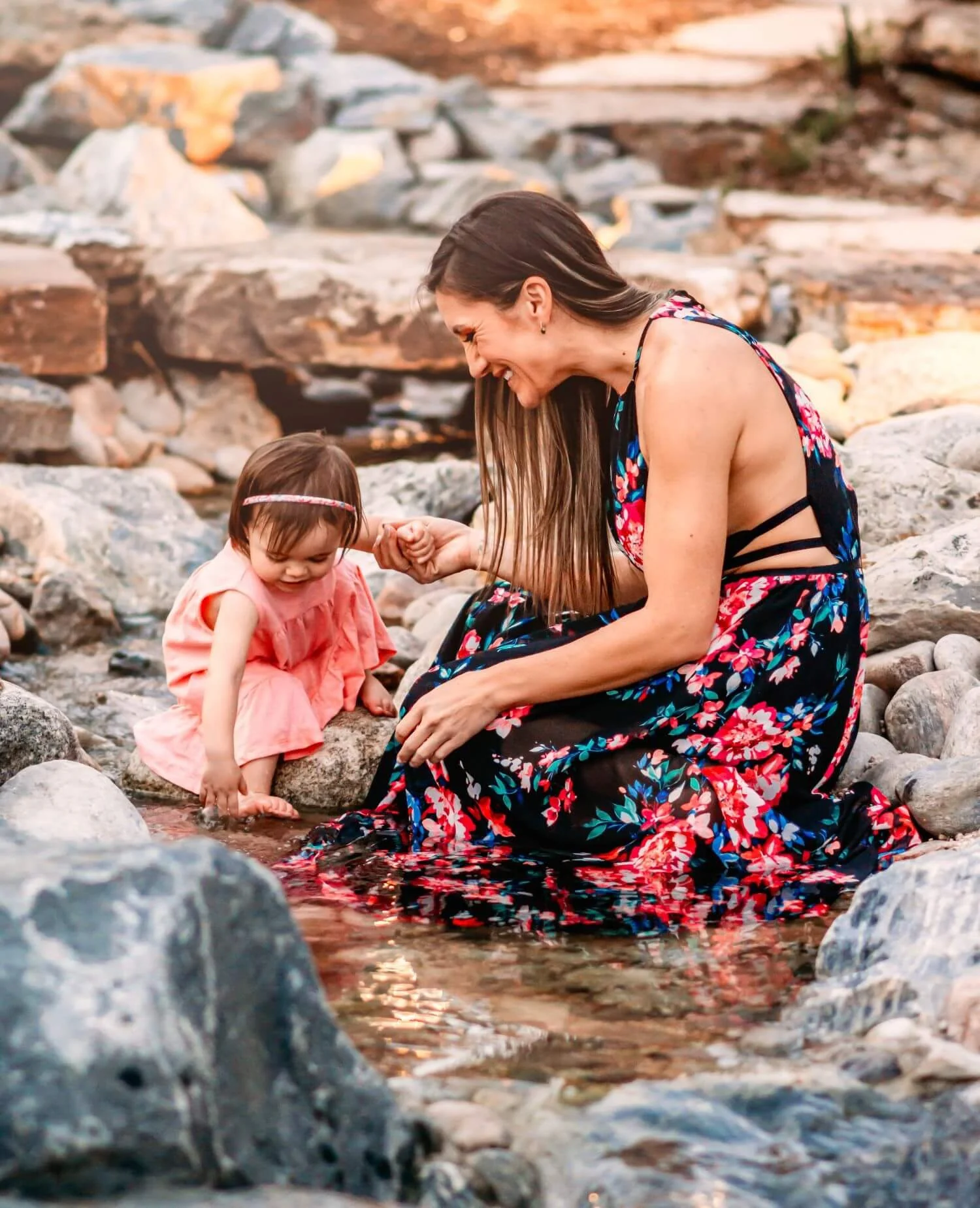 A woman and a young girl sit on rocks by a small stream, smiling and holding hands, surrounded by rocks and natural outdoor scenery.