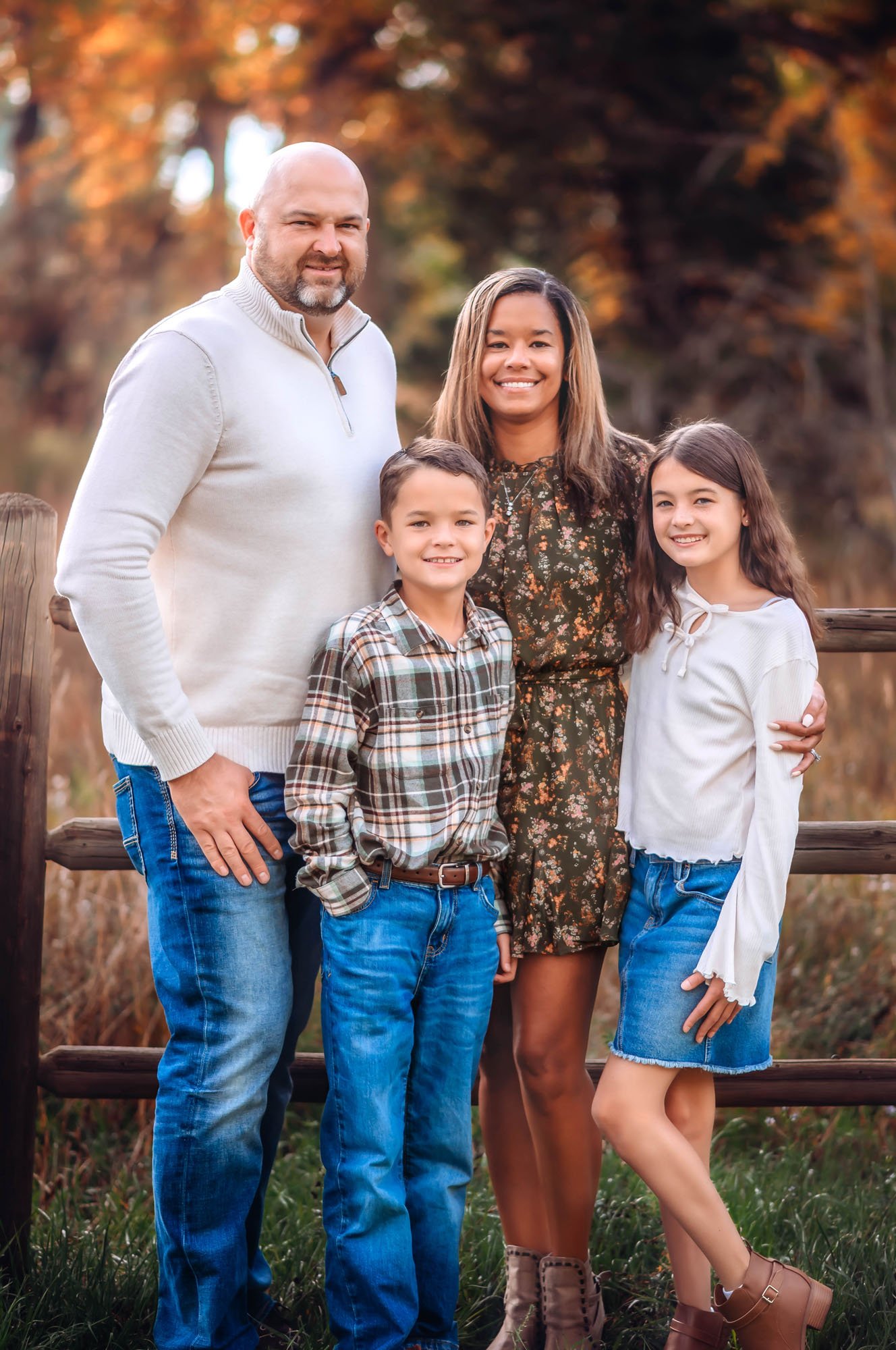 Family of five outdoors during autumn with trees in background. Adults and children smiling, wearing casual clothing and boots.