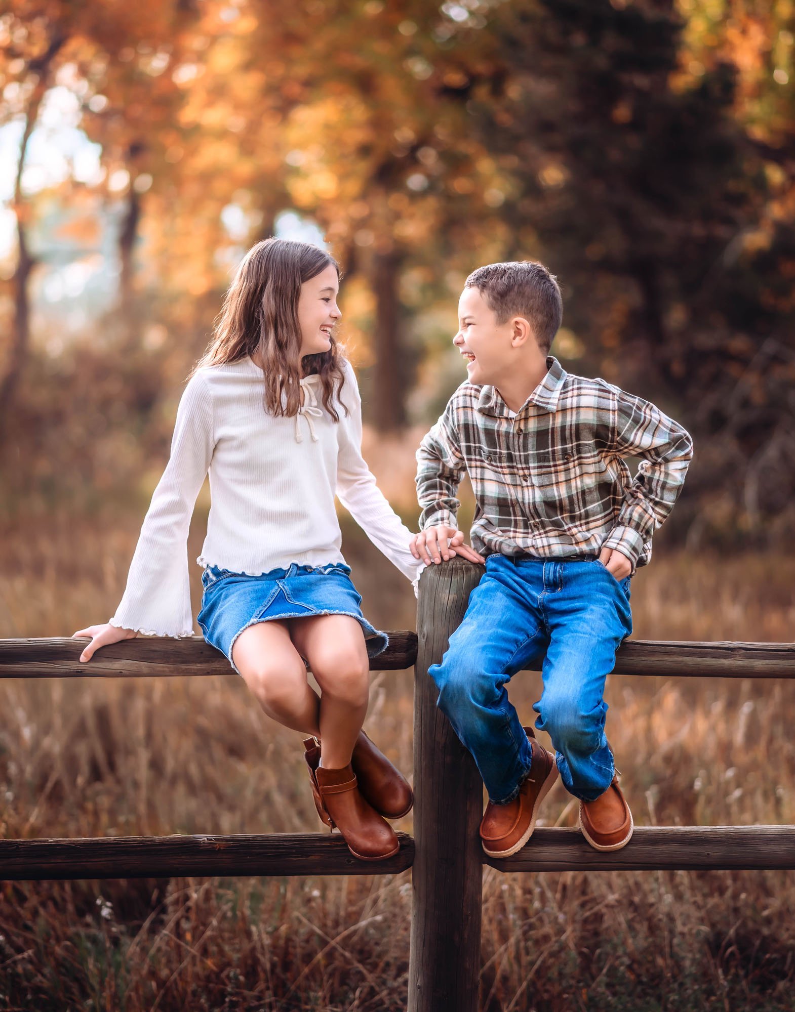 Two children, a girl and a boy, sitting on a wooden fence outdoors during autumn, holding hands and smiling at each other.