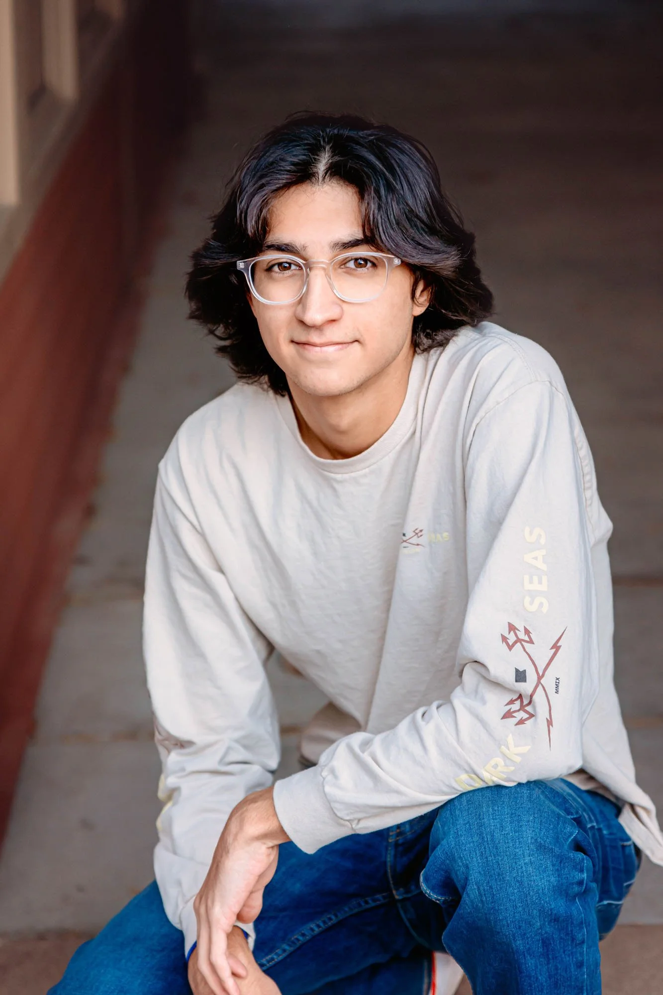 A young man with medium-length dark hair, glasses, and light skin, wearing a light beige sweatshirt with graphic design on the sleeves and blue jeans, sitting on stairs outdoors.