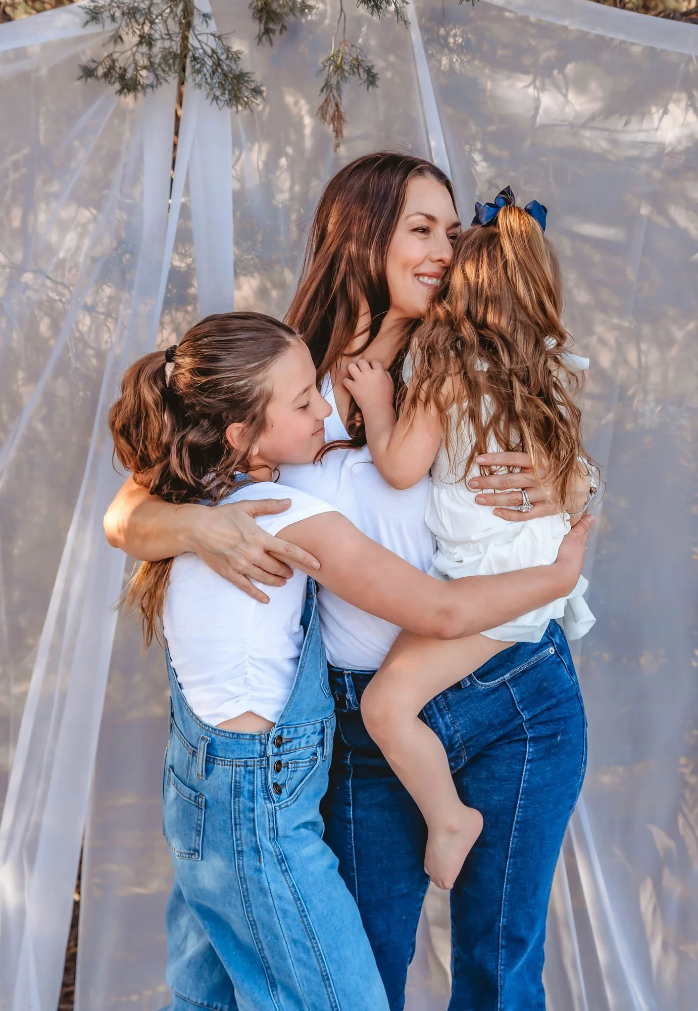 A woman and two young girls hugging each other outdoors, with a white fabric or tent in the background.