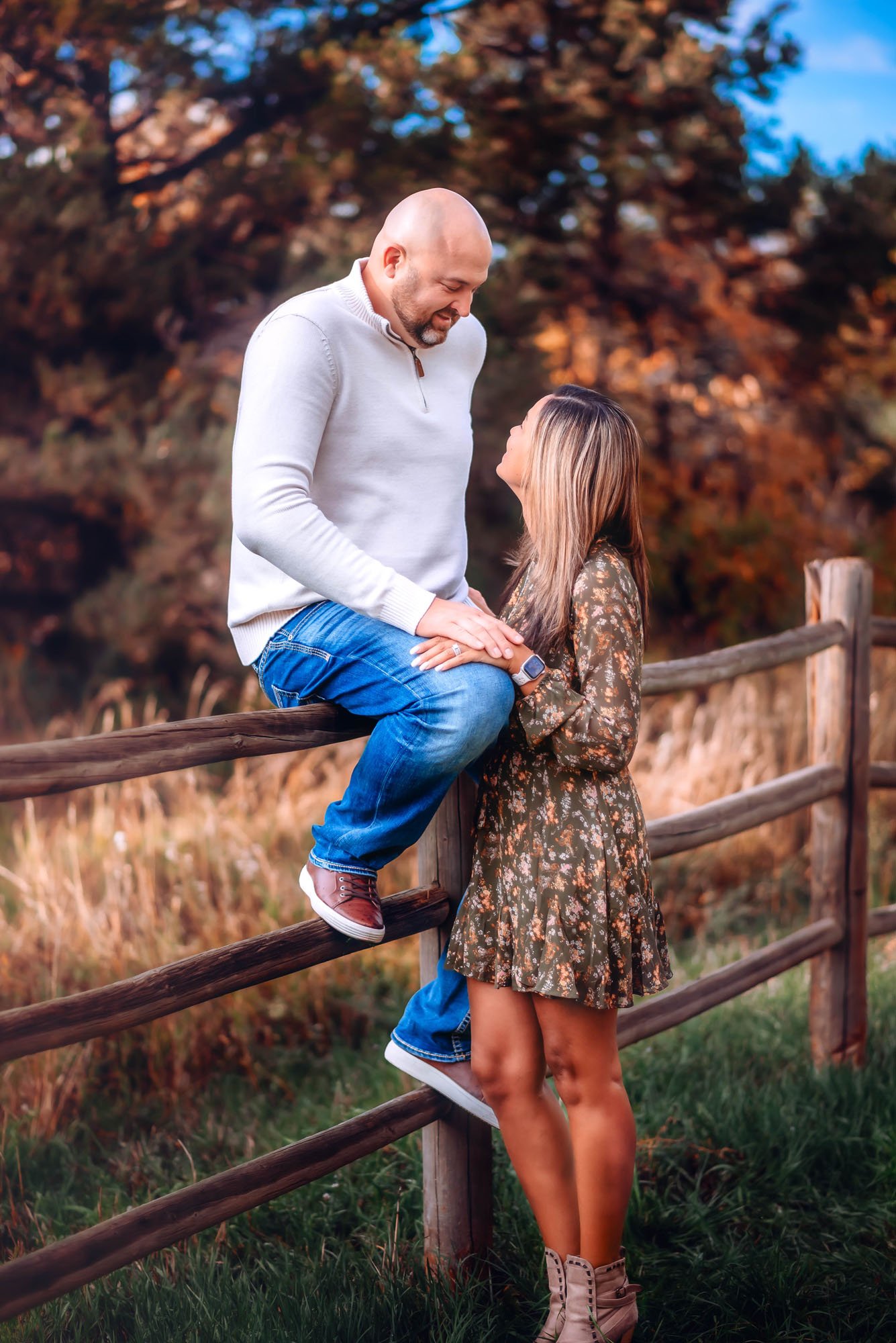 A couple standing outdoors near a wooden fence, during autumn with trees in the background. The man is seated on the fence, looking down at the woman who is standing and looking up at him. They are holding hands and holding each other. The man is bal