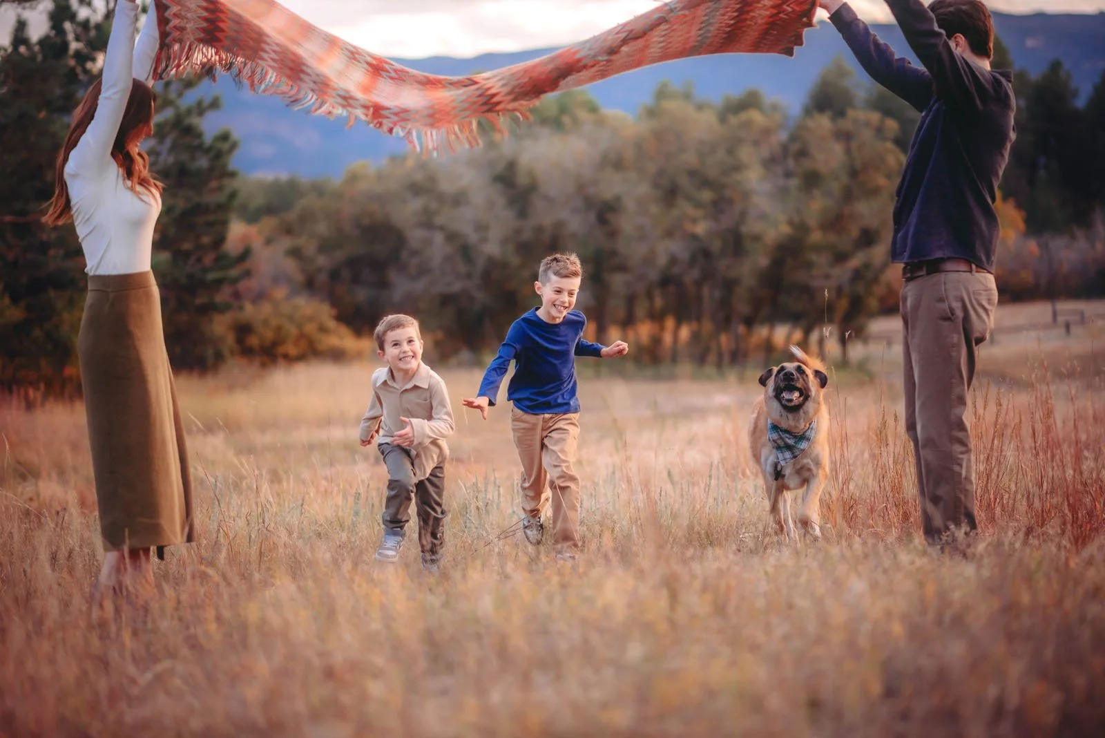 A family of four and a dog in a field during autumn. The father and mother are holding up a blanket while two young boys run and play, with trees and mountains in the background.