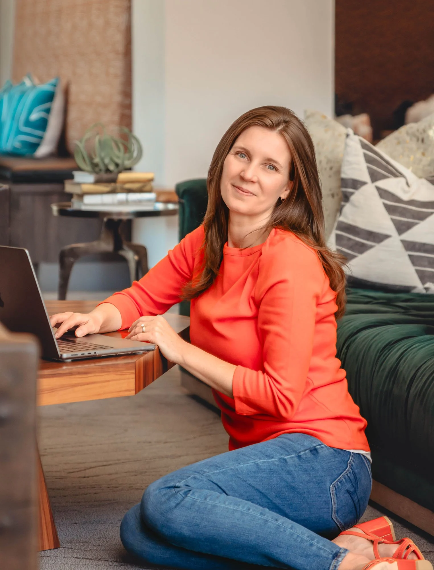 A woman with long brown hair, wearing an orange top, blue jeans, and orange sandals, is sitting on the floor with her legs crossed, working on a laptop on the desk in front of her. She is looking at the camera and smiling. The background features a c