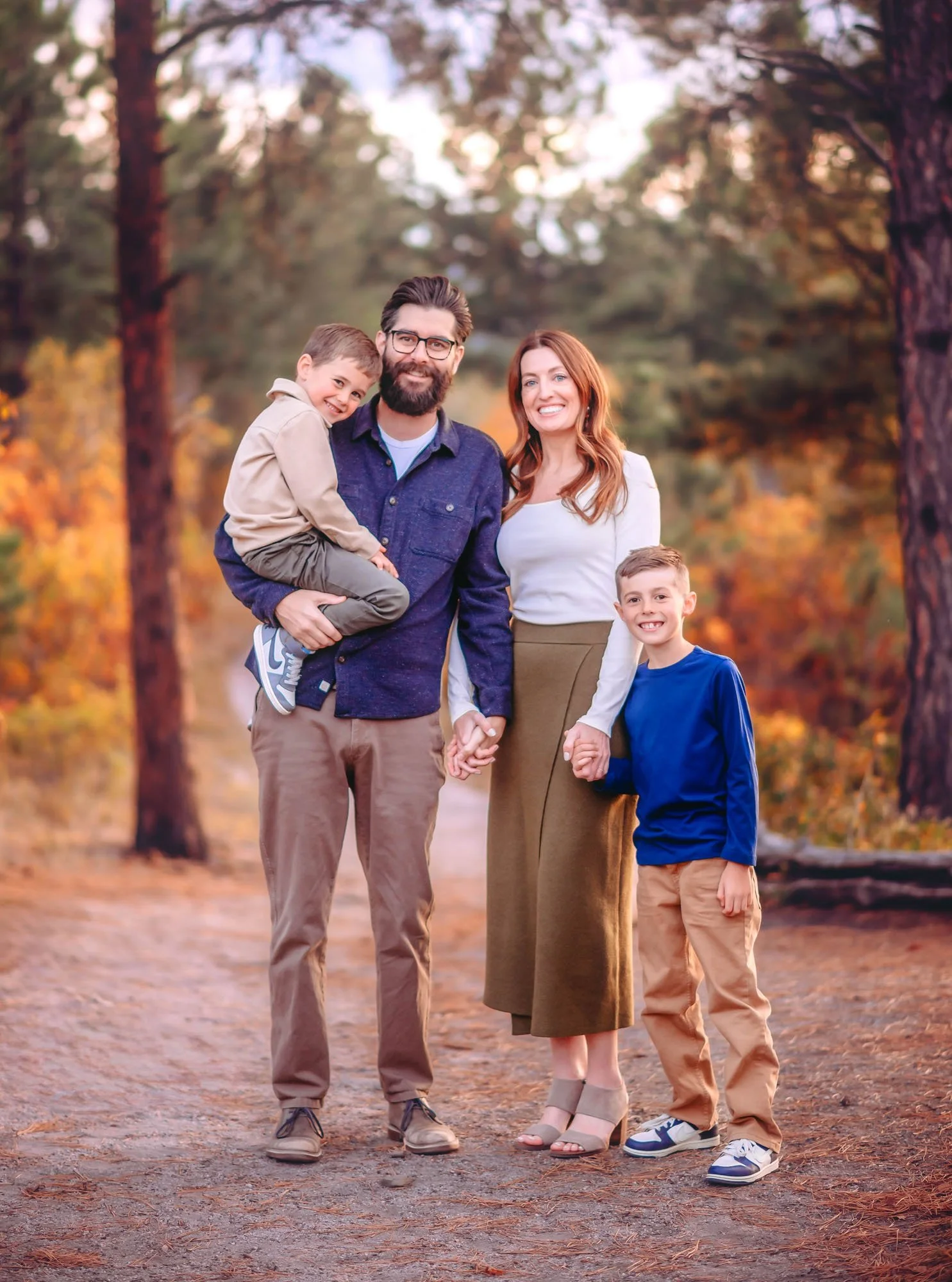 A happy family of four standing outdoors on a dirt path surrounded by trees with fall foliage, smiling at the camera. The father is holding a young boy, and the mother is next to the older son.