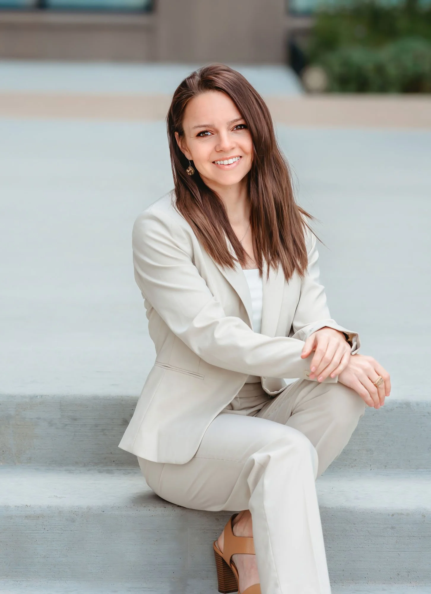 A woman with long brown hair, wearing a beige suit and matching pants, is sitting on the ground outside on a concrete surface near water. She has an earring in one ear and is smiling at the camera, with her hands resting on her knee. She is wearing t