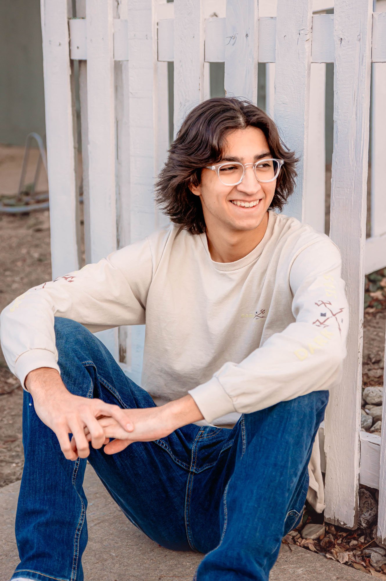 A young man with long wavy hair and glasses sitting outside near a white wooden fence, smiling, wearing a beige long-sleeve shirt and blue jeans.