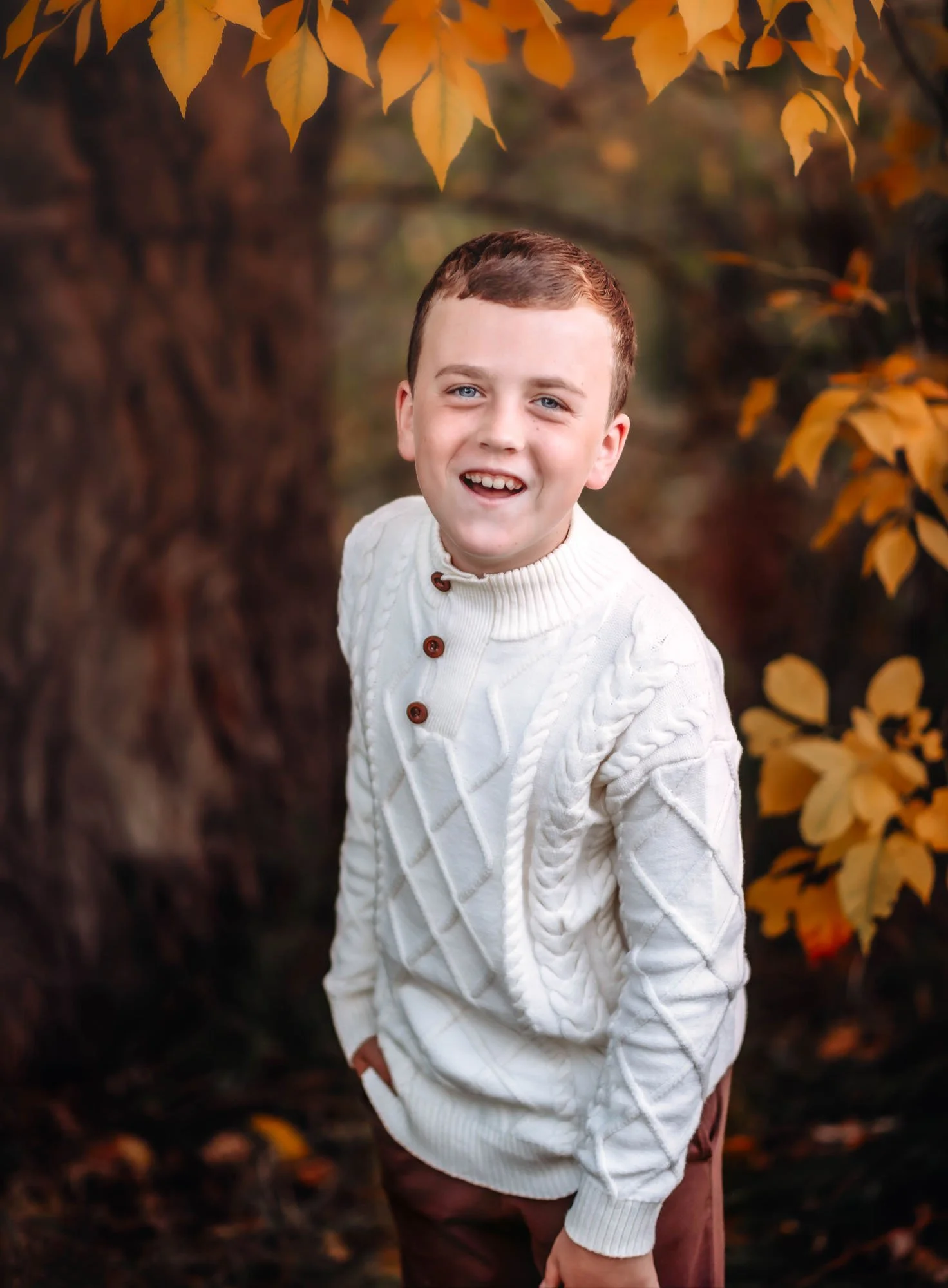 A young boy with short brown hair and blue eyes smiling outdoors in front of autumn-colored leaves, wearing a cream-colored cable knit sweater with brown buttons.