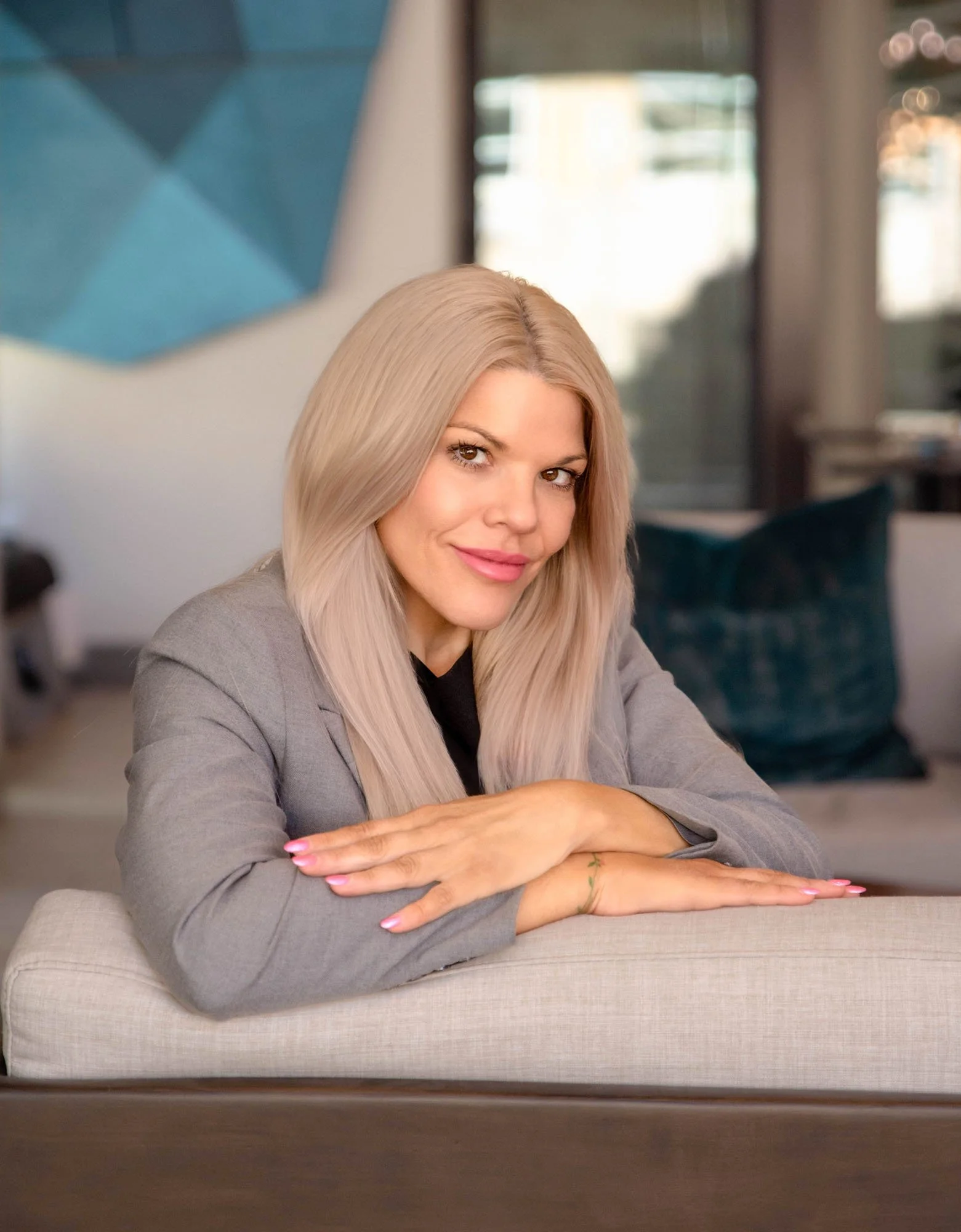 A woman with long, platinum blonde hair and tan eyes sitting on a beige couch in a bright room, wearing a gray blazer and smiling at the camera.