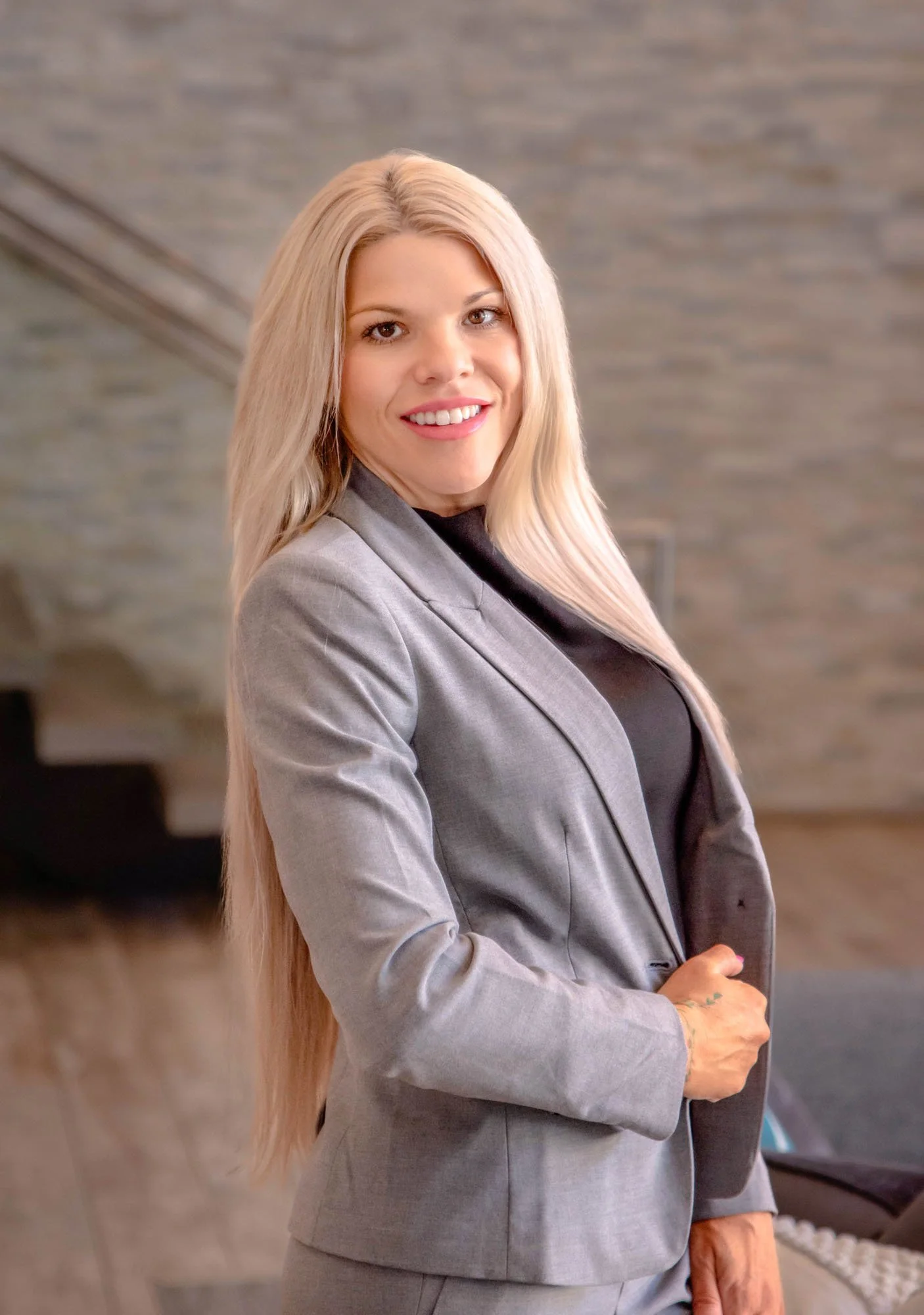 A woman with long blonde hair, wearing a gray blazer and black top, smiling and looking into the camera in an indoor setting with a brick wall background.