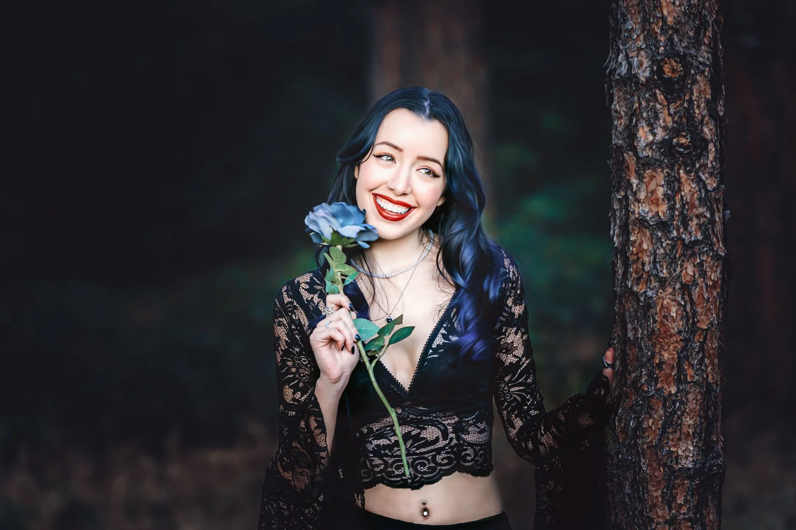 A woman with long dark blue hair holding a purple flower in a forest with tall trees.