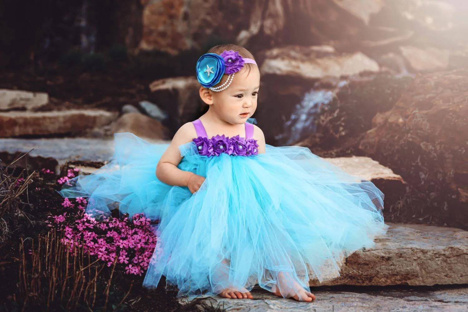 A young girl dressed in a blue tulle dress with purple floral embellishments on the bodice and strap, sitting barefoot on rocks outdoors near a small stream, with pink flowers and brown rocks surrounding her.