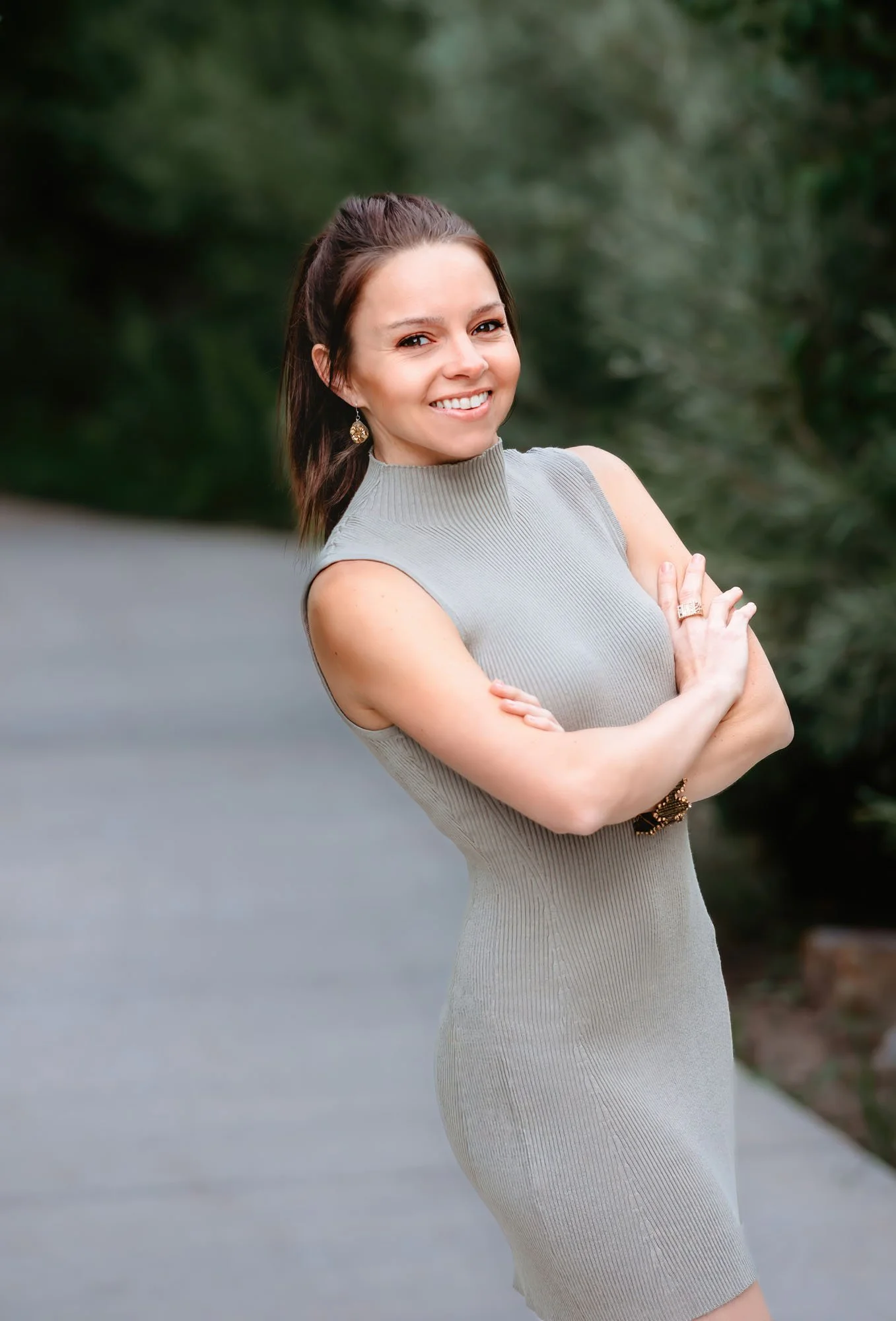 Smiling woman with brown hair in a half-up half-down style, wearing a sleeveless gray ribbed dress and gold jewelry, crossing arms outdoors with blurred greenery background.