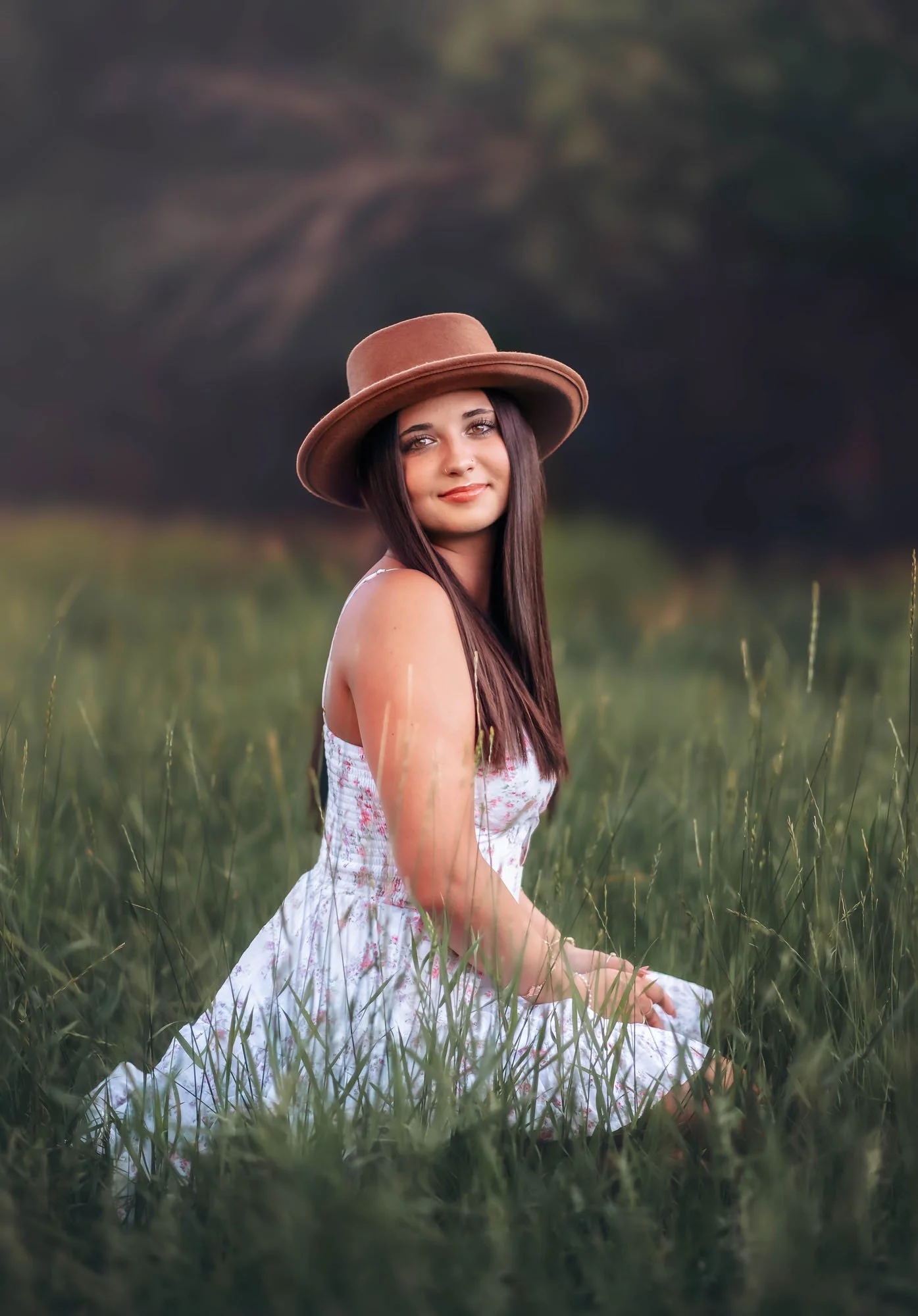 A young woman with long dark hair sitting in green grass, wearing a wide-brimmed brown hat and a pink and white floral dress, smiling and looking at the camera in an outdoor setting with blurred trees in the background.