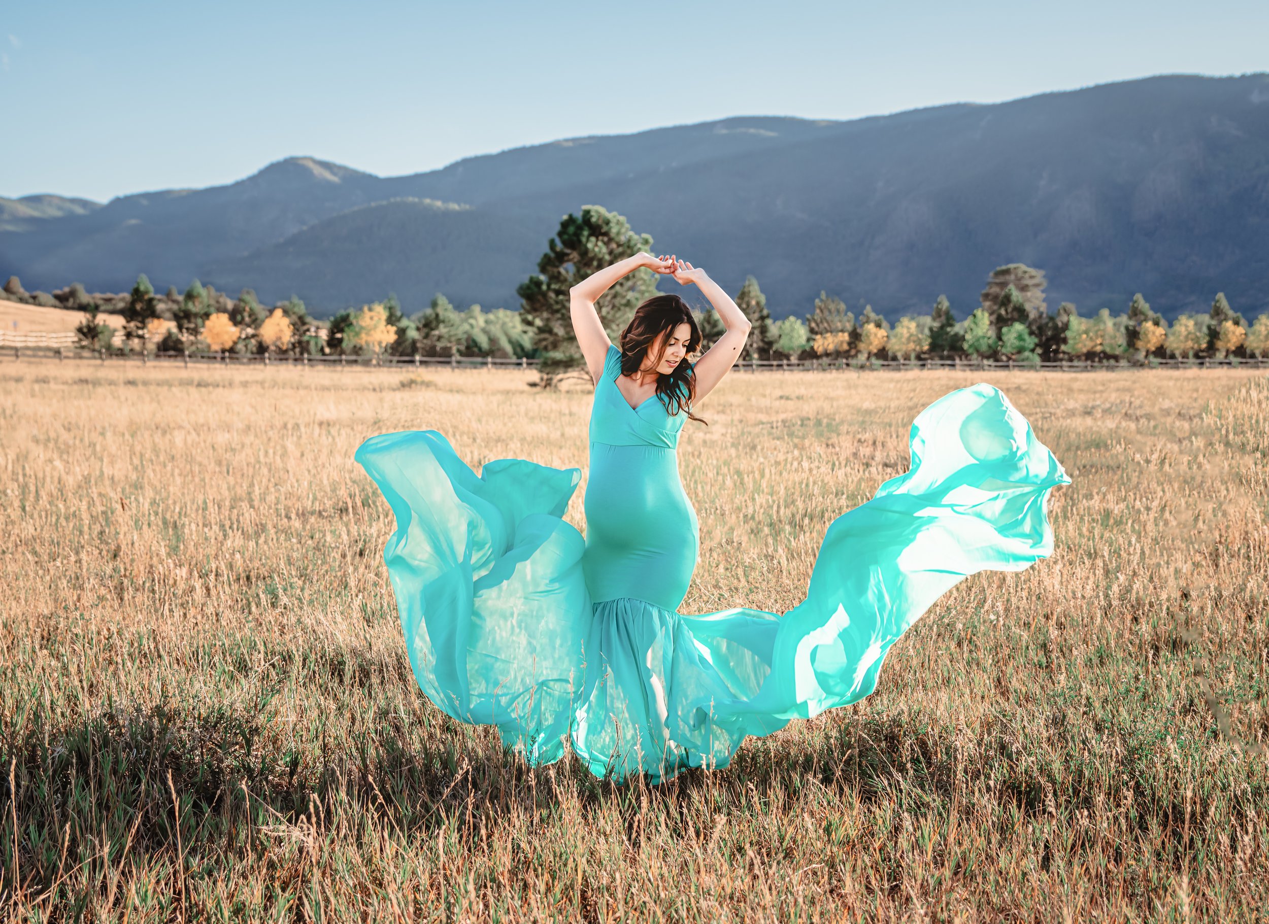A woman in a turquoise dress dancing in a field with mountains in the background, with flowing fabric around her.