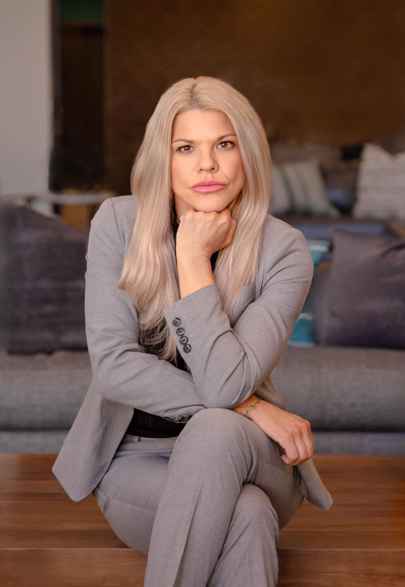 A woman with long blonde hair in a light gray business suit sits on a wooden bench in a modern room with a dark brown wooden accent wall and a bed with pillows behind her. She is resting her chin on her hand and looking directly at the camera.