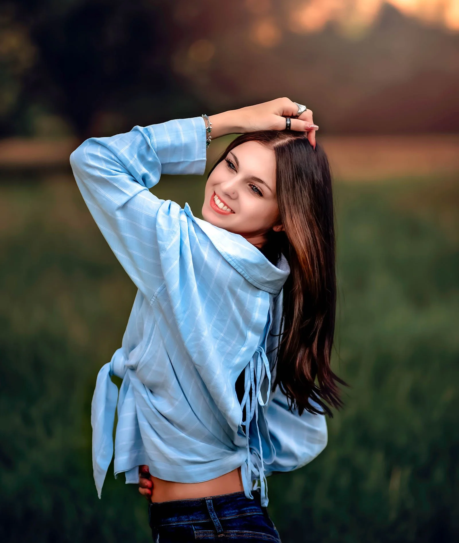 A woman with long brown hair posing outdoors in a blue checkered shirt at sunset.