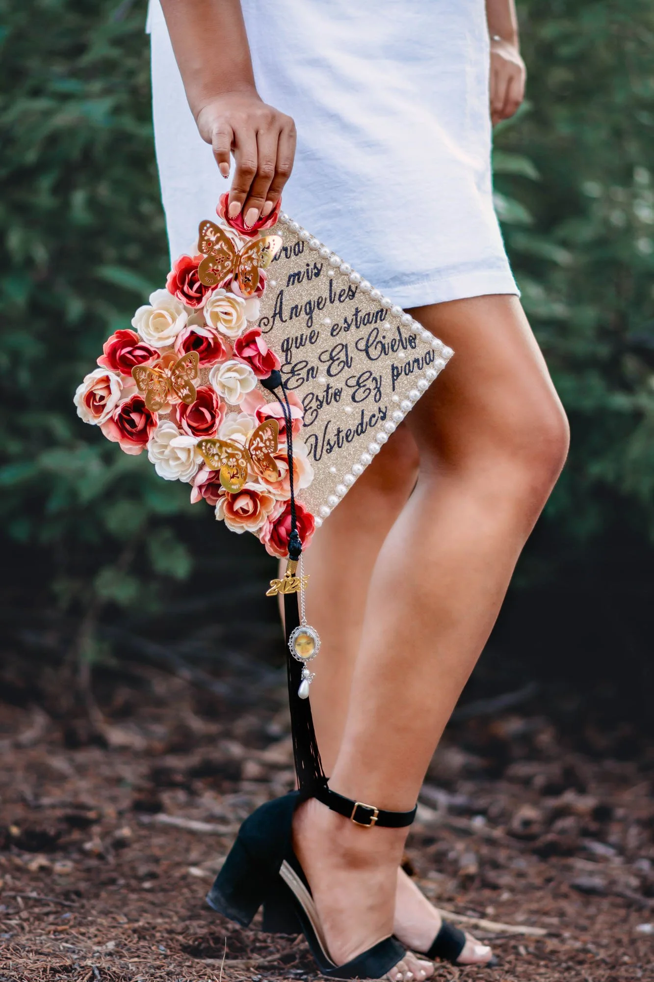 Close-up of a woman wearing a white dress and high heels, holding a decorated graduation cap with flowers, butterflies, and charms, on a forest floor.