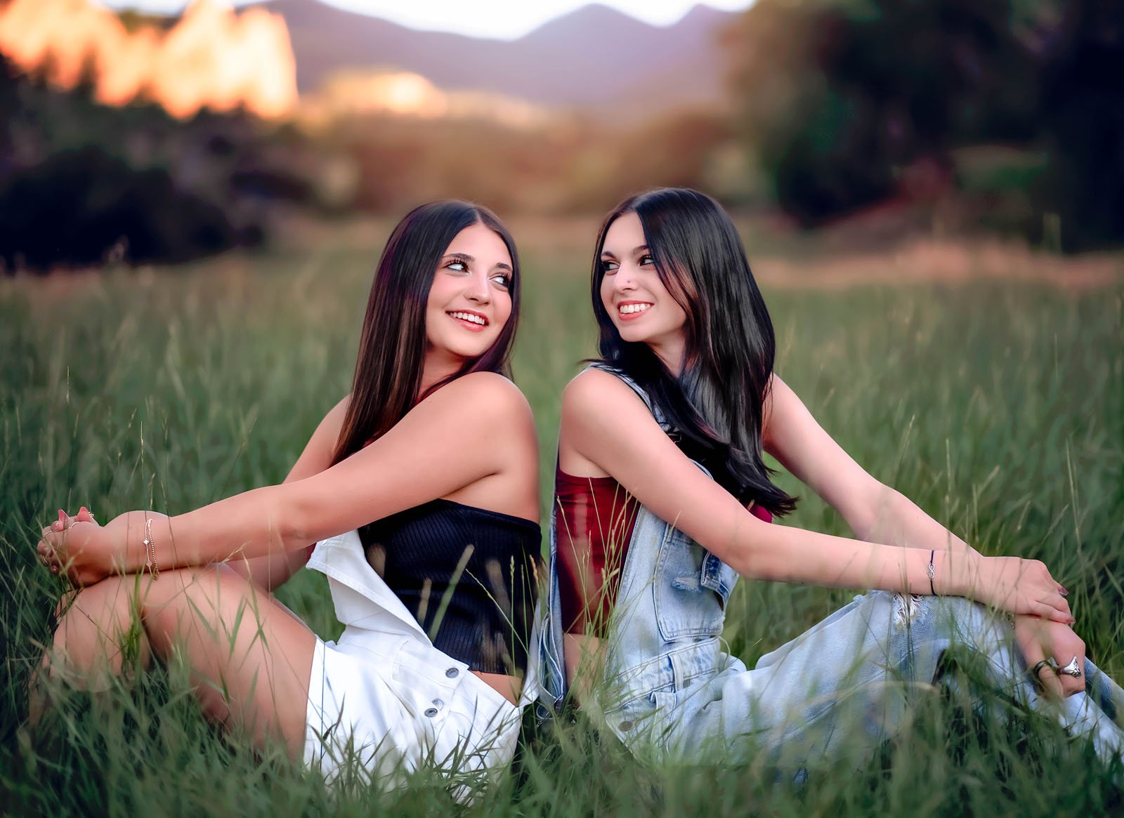 Two young women sitting in a grassy field, smiling at each other, with blurred trees and sunset in the background.
