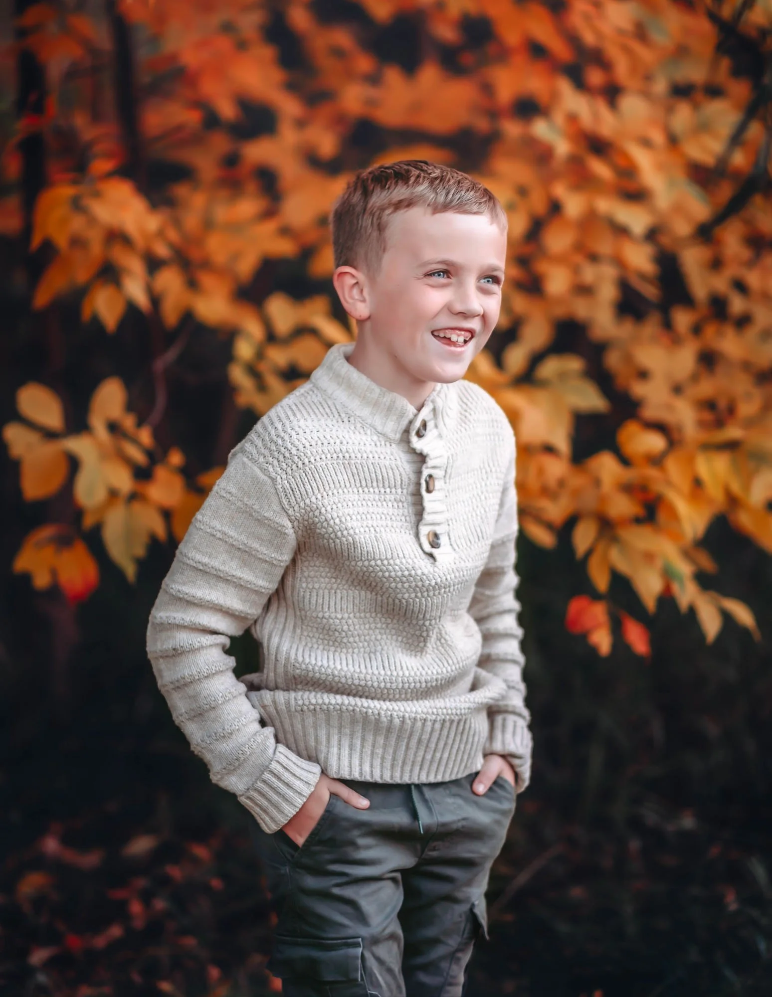 A young boy with short reddish hair and blue eyes smiling while standing outdoors among orange autumn leaves. He is wearing a cream-colored knit sweater and gray cargo pants with his hands in his pockets.