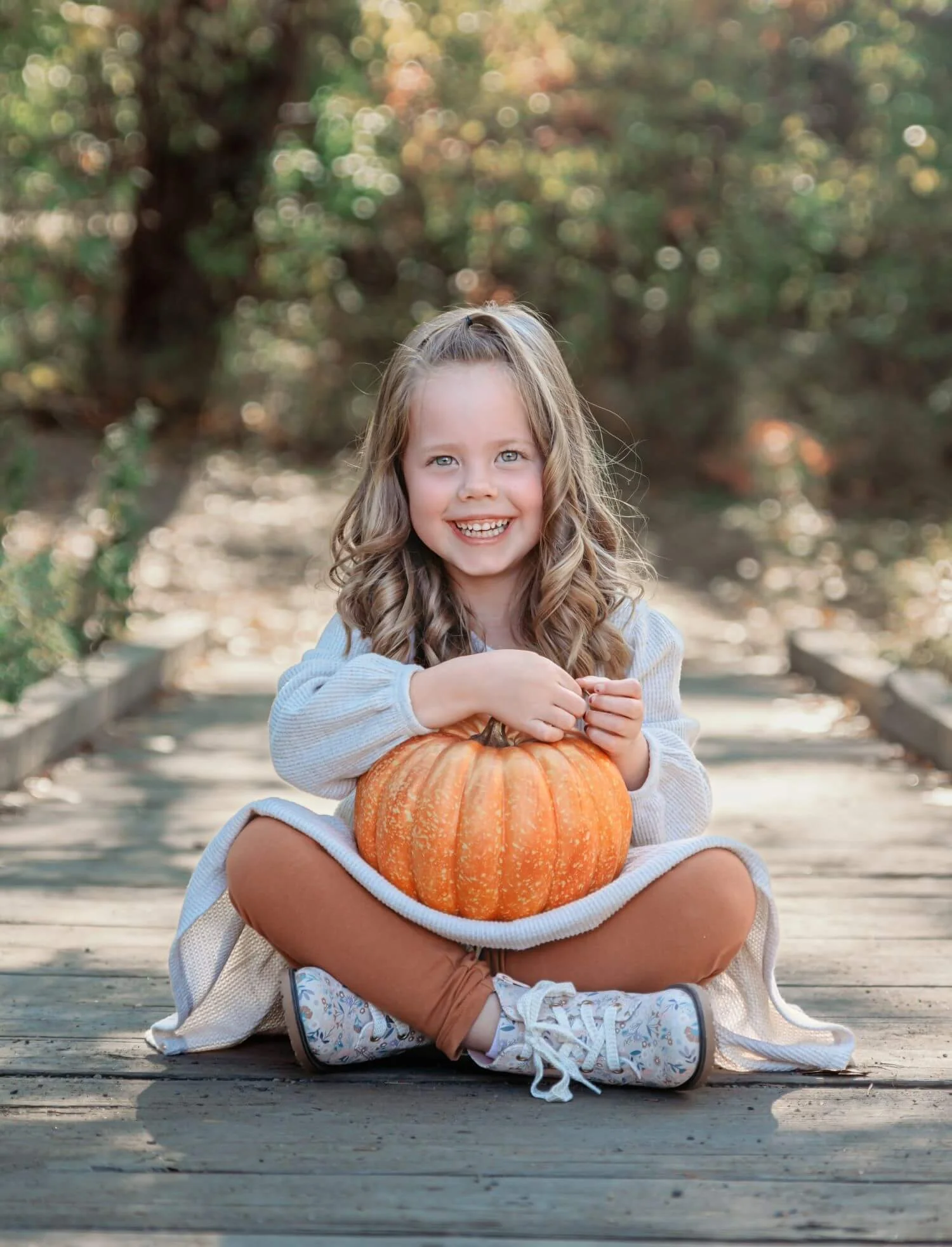 A young girl sitting cross-legged on a wooden walkway outdoors, smiling, holding a large orange pumpkin in her lap, with autumn trees in the background.