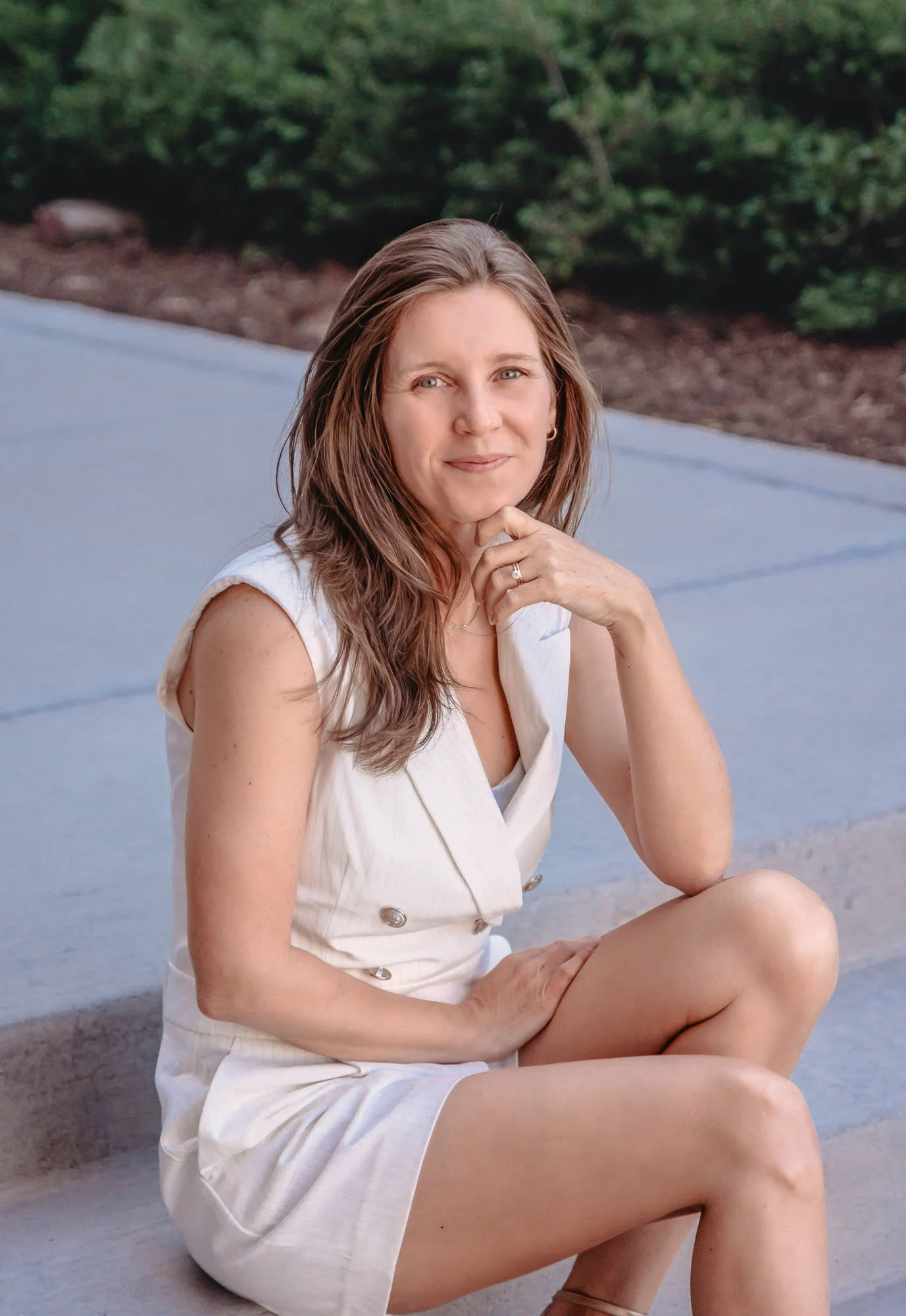 A woman sitting outdoors on a bench, with a background of green trees, wearing a sleeveless white dress and resting her chin on her hand.