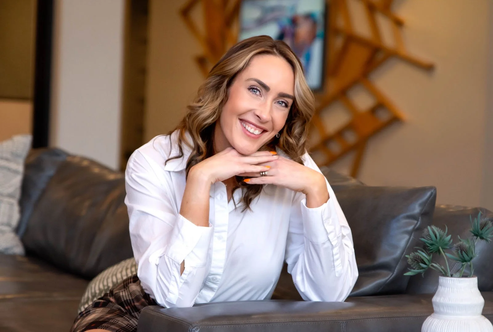A woman with wavy light brown hair and blue eyes smiling while sitting on a couch in a living room.