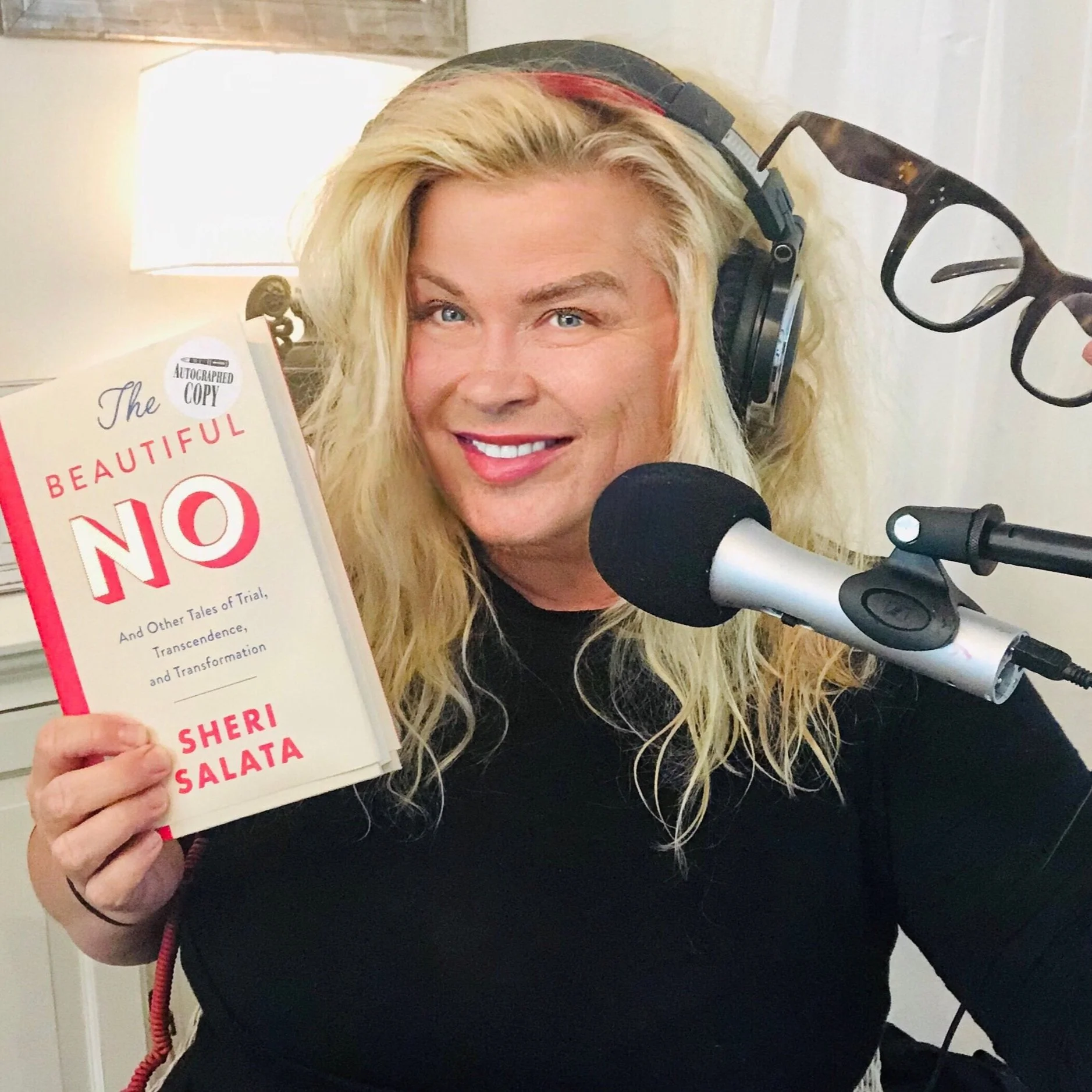 Woman holding a pair of glasses in one hand and a book in the other. In studio with a microphone in front of her.