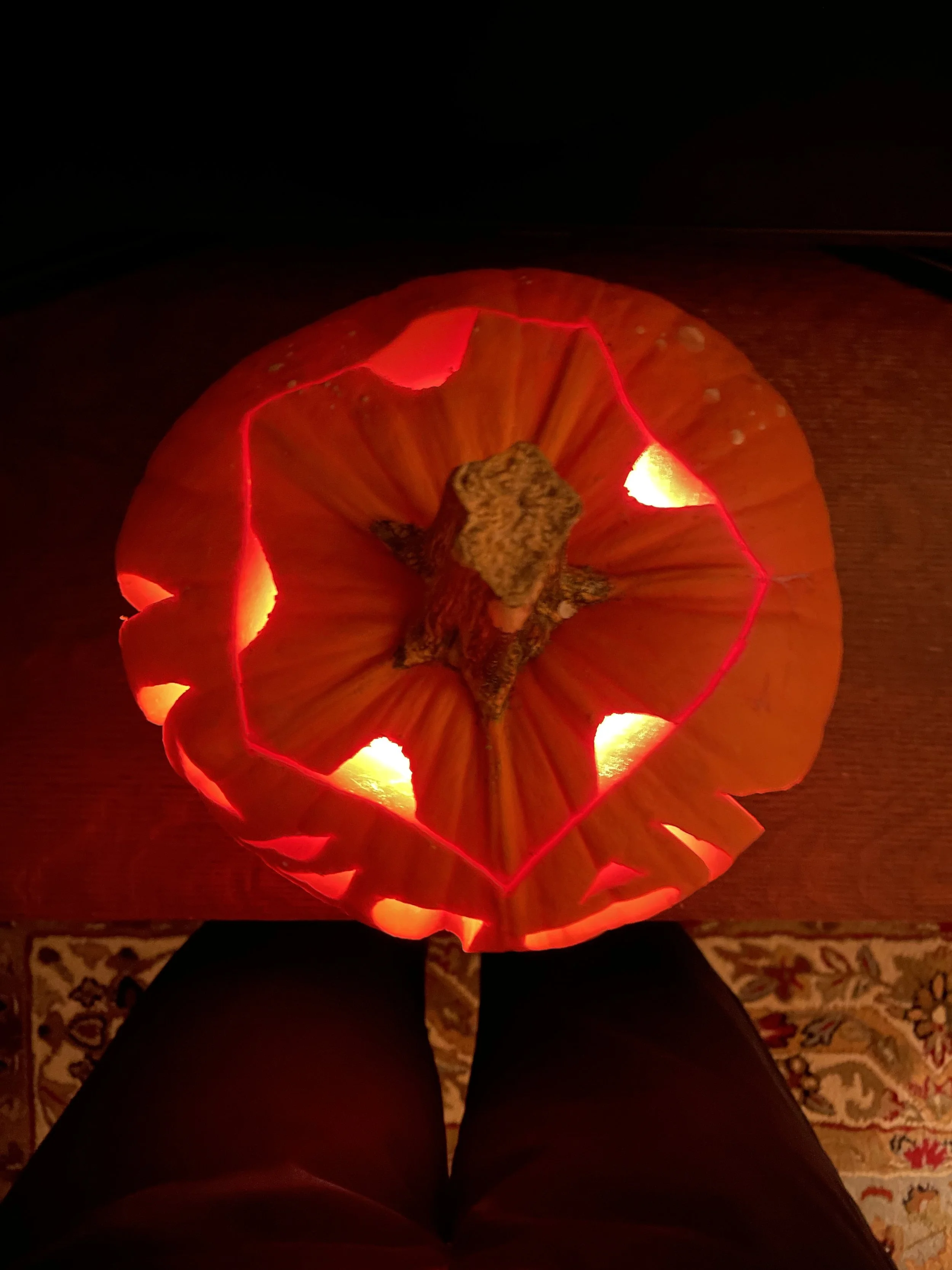 A photo of a carved pumpkin in the dark. Inside it is illuminated. Shot from above, the top features a pentagon shape with small semi-circle cutouts. 