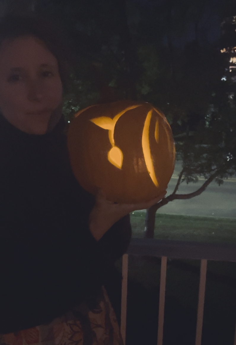 A photo of a carved pumpkin in the dark, held by a woman. It is illuminated. This side of the pumpkin shows a sprout dropping with a side leaf and budding leaf