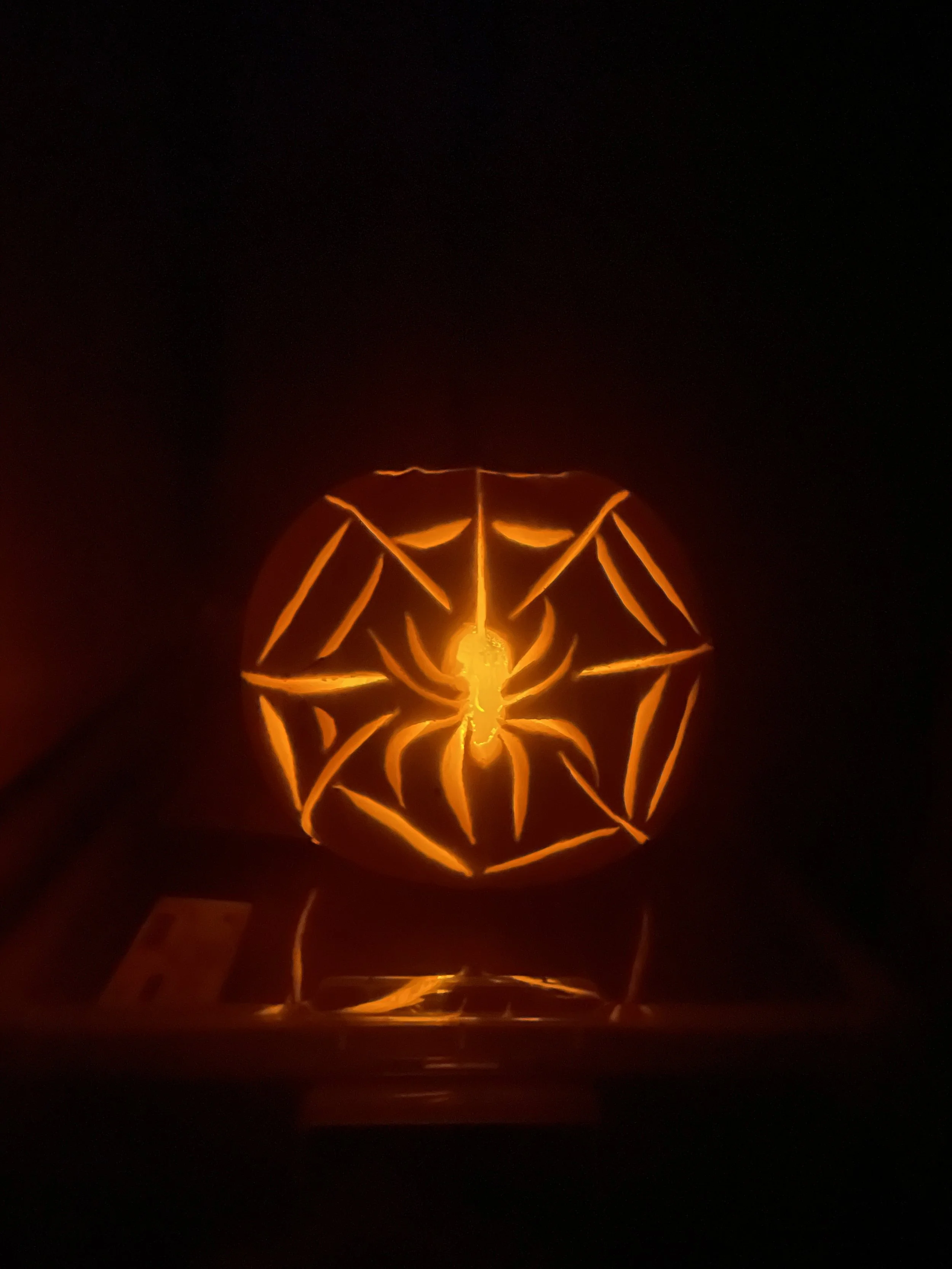 A photo of a carved pumpkin in the dark. Inside it is illuminated. It features a spider on a web.