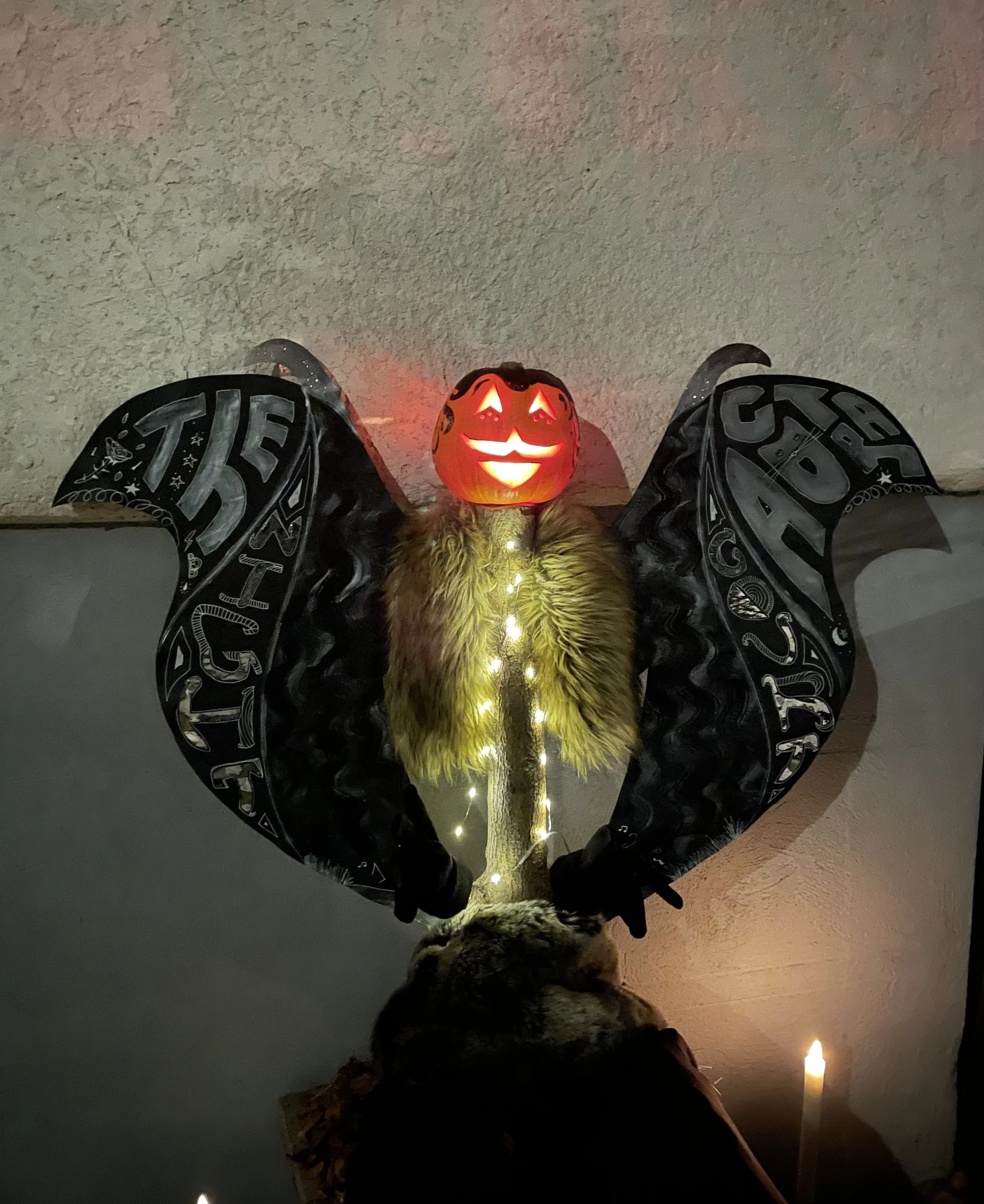 A photo of a carved pumpkin in the dark. Inside it is illuminated. The pumpkin sits atop a tree stump and has illustrated black wings behind it. It wears a fauc fur scarf and is mounted.