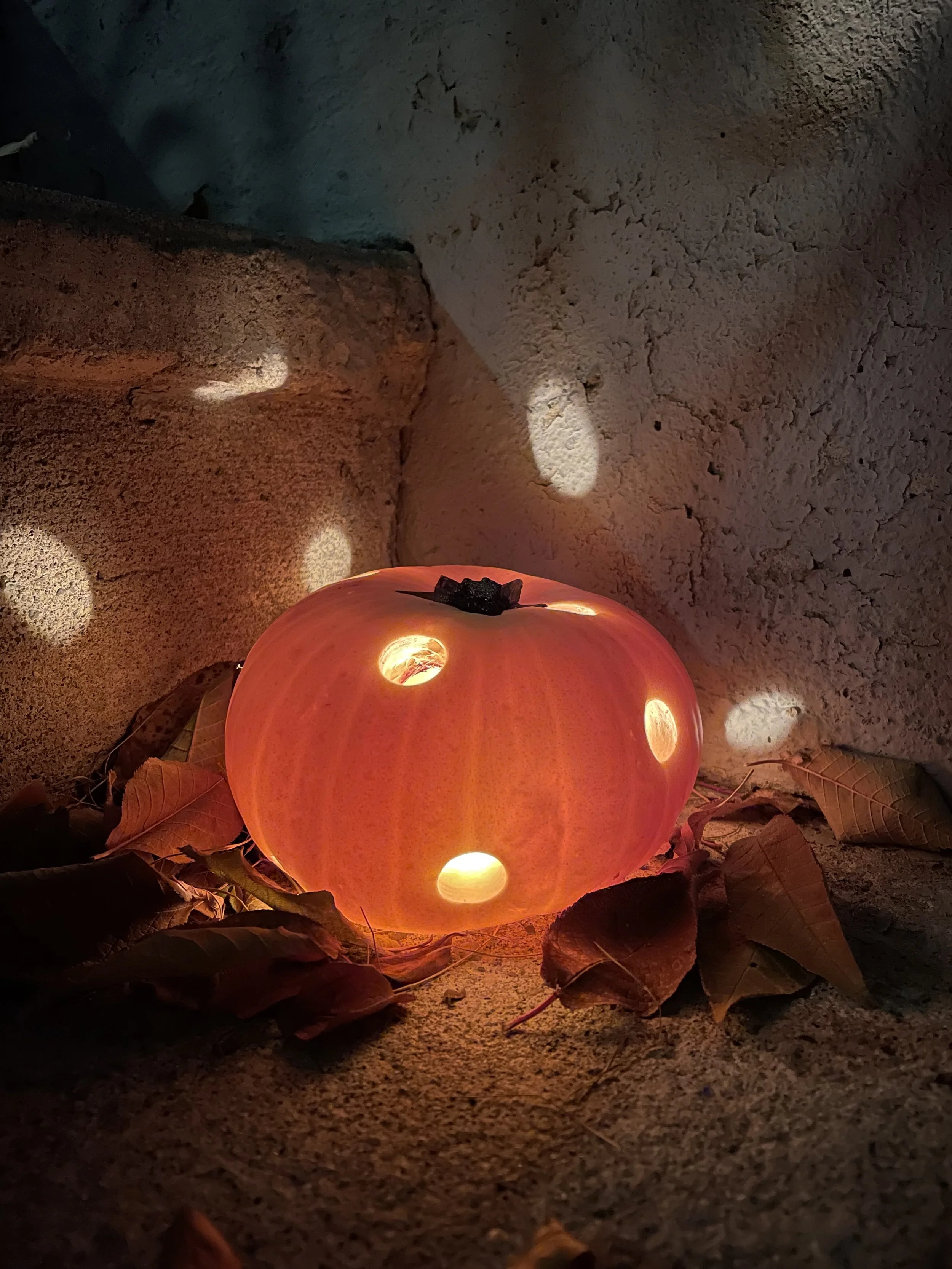 A photo of a carved pumpkin in the dark. Inside it is illuminated. It is carved to be a like a mushroom top. With big, even holes made by a drill. It looks like a disco pumpkin. It sits on leaves