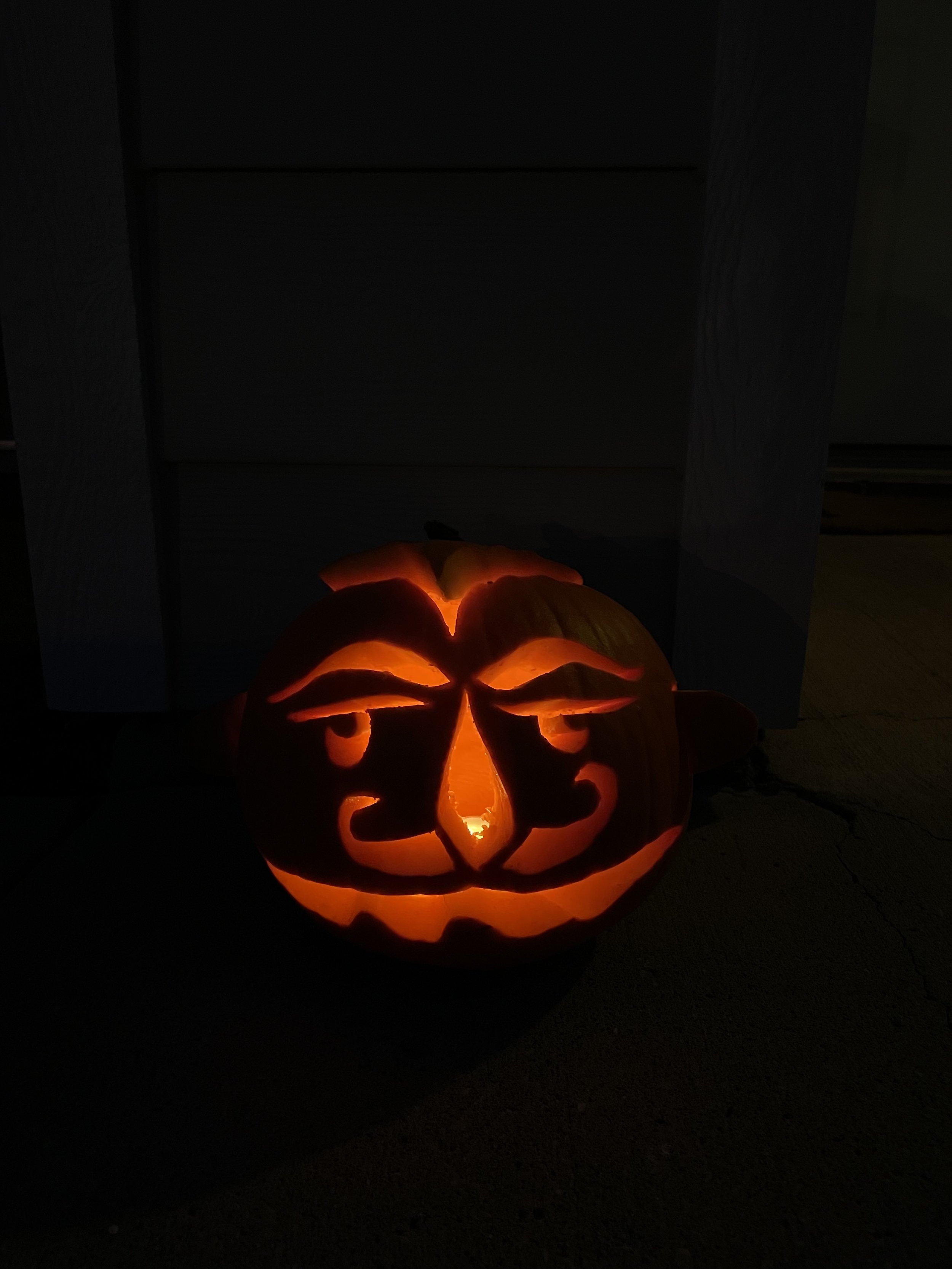 A photo of a carved pumpkin in the dark. Inside it is illuminated. Inspired by an elder Greek woman's deceased husband, this pumpkin is of a stylized man's face. Curled moustache, kind eyes. 