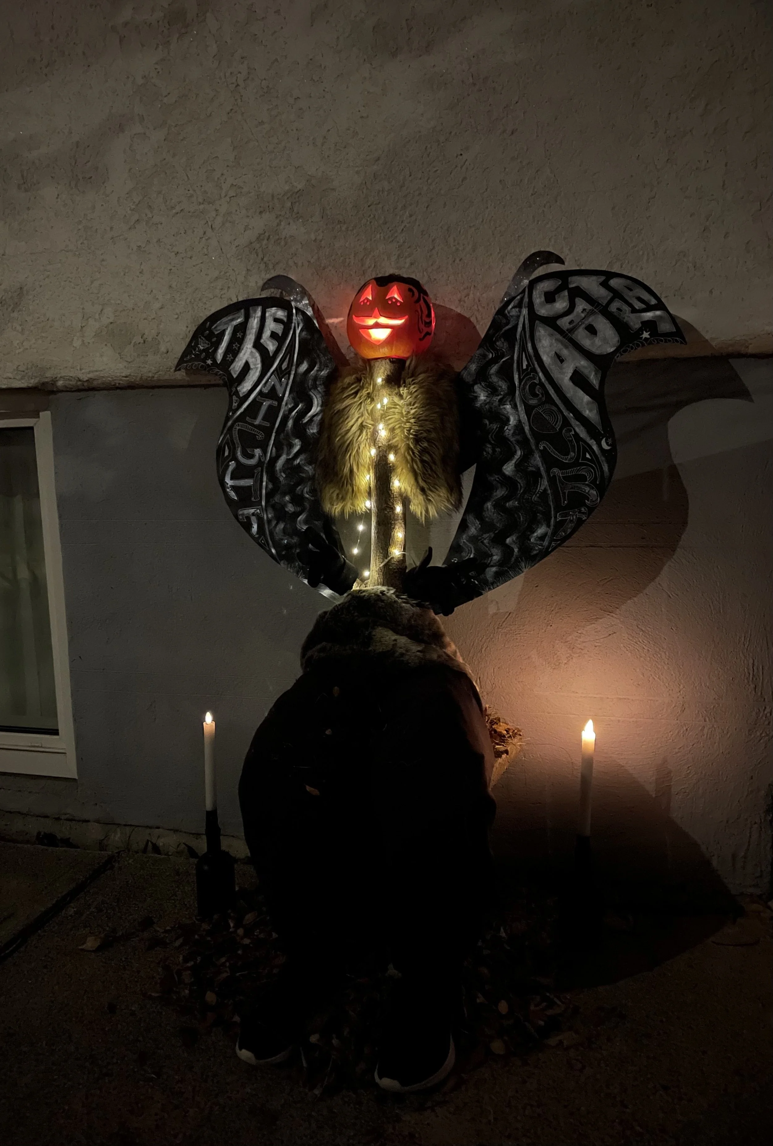 A photo of a carved pumpkin in the dark. Inside it is illuminated. A full install of a scarecrow-like pumpkin with faux candlesticks on either side. It stis atop a tree stump and has a string of light going down the stump. It has decorated wings.